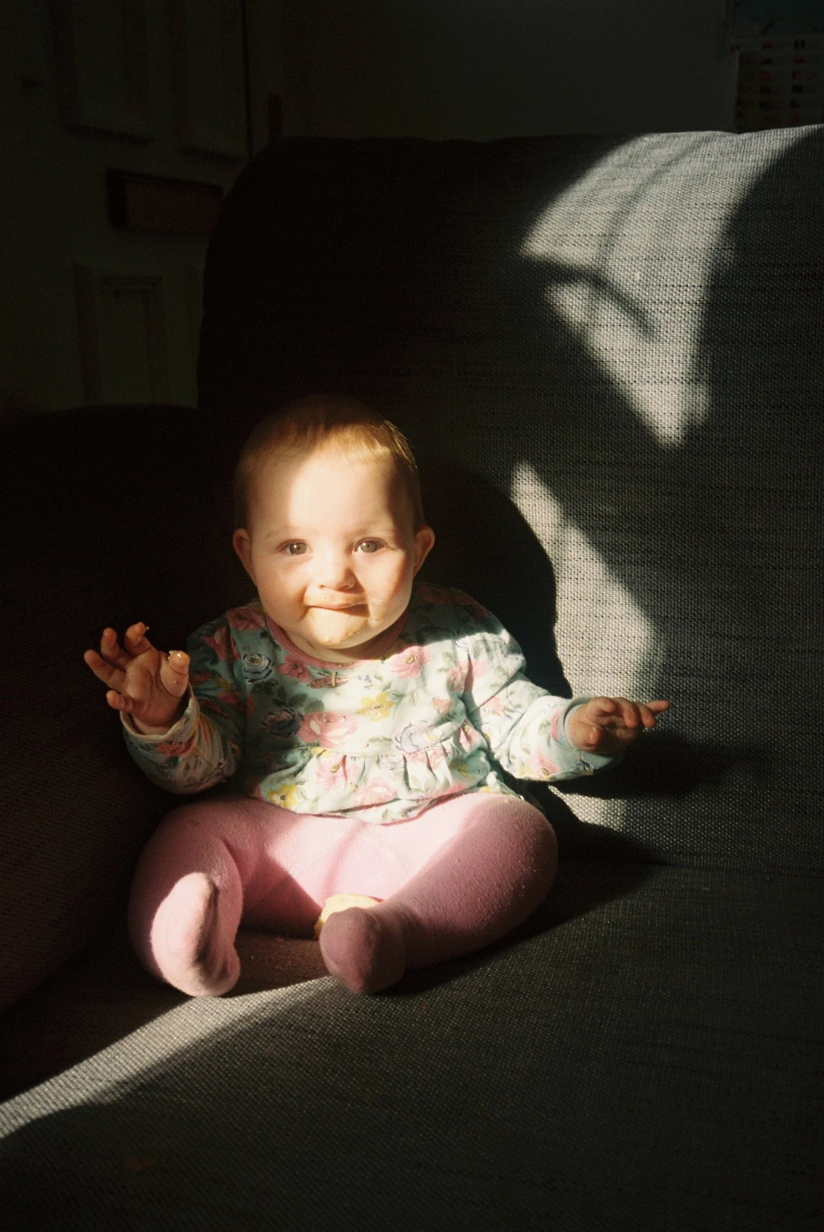 A baby sitting on a couch in sunlight, wearing a floral shirt and pink leggings.