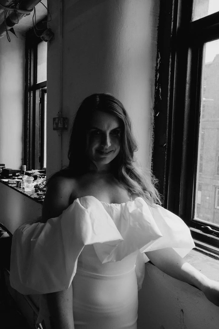 Black and white photo of a woman in an off-shoulder Jessica Bennet dress standing by a window, with soft lighting and shadow effects.