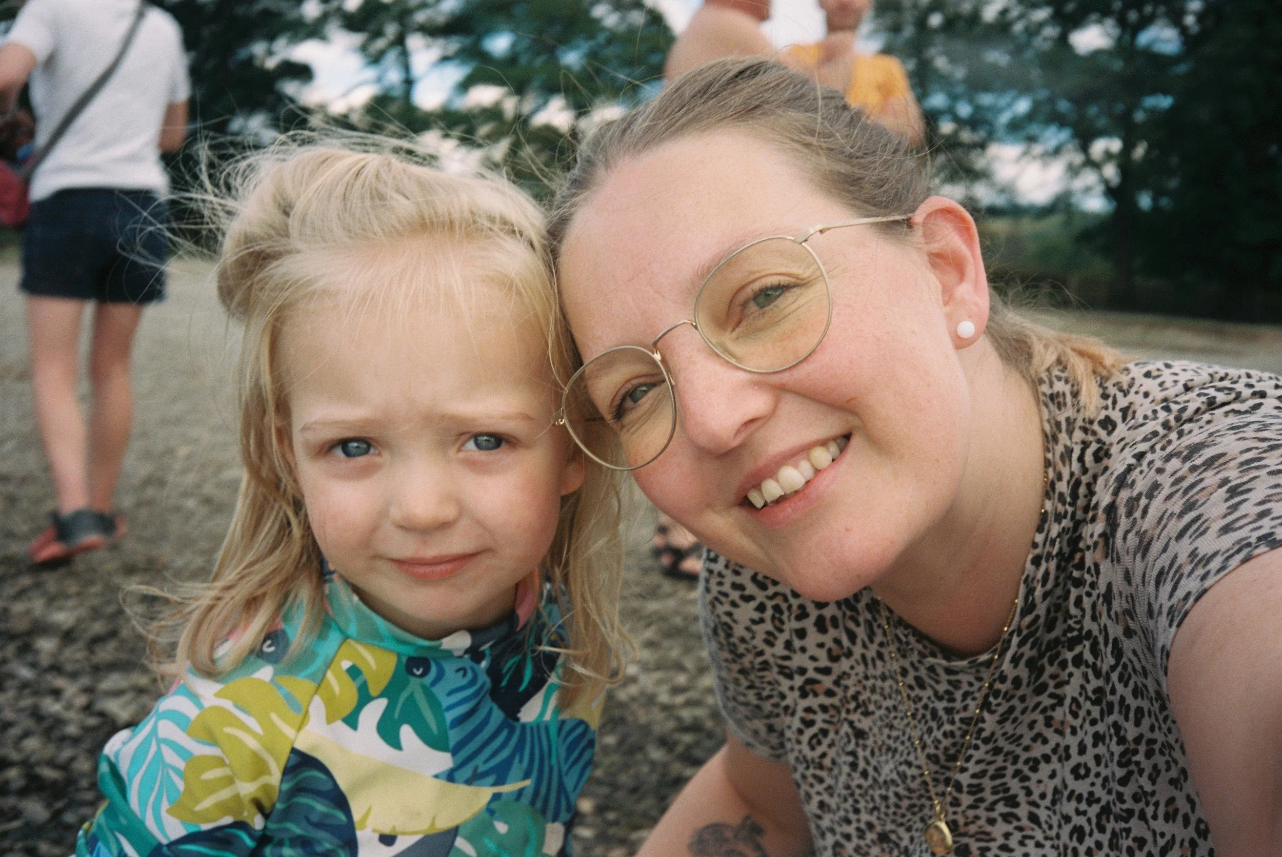 Adult woman and child outdoors smiling close-up, with trees and people in the background.