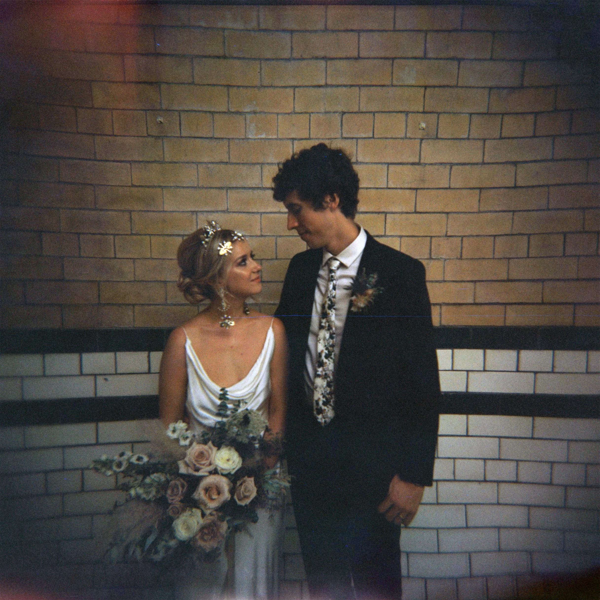 Becca & Callum stand with their backs to the wall of Victoria Baths in Manchester. Becca has statement earrings and headpiece on from Tilly Thomas Lux and is wearing a Ghost Orchid wedding dress and Callum a black suit with flowery tie.