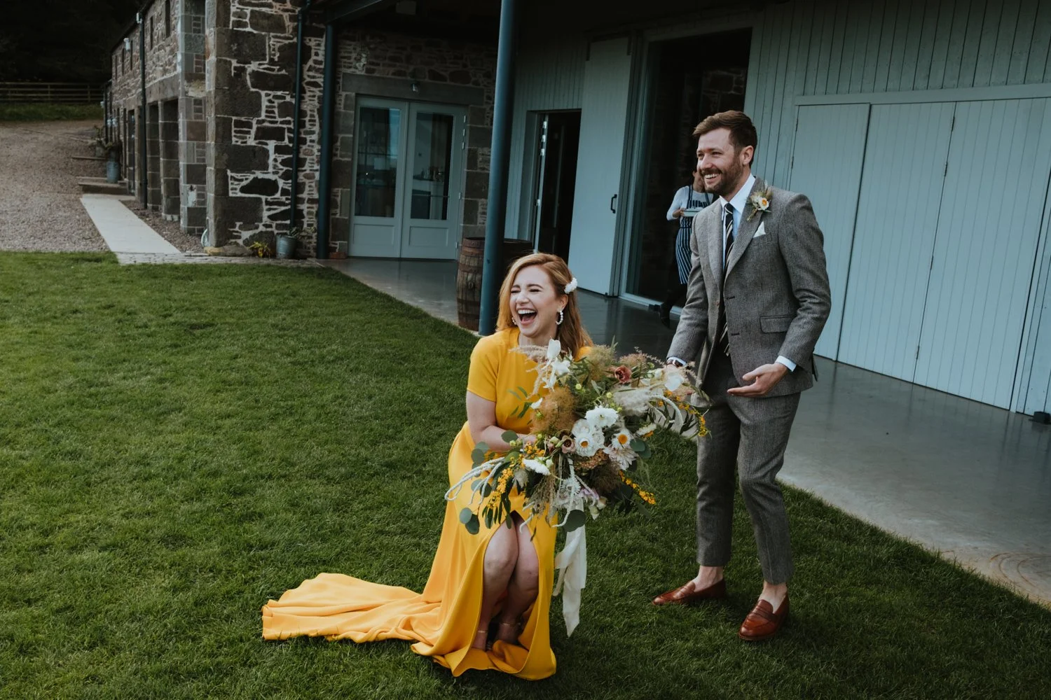 A woman in a yellow dress holding a bouquet and laughing next to a man in a gray suit, standing on a grassy area by a rustic building.