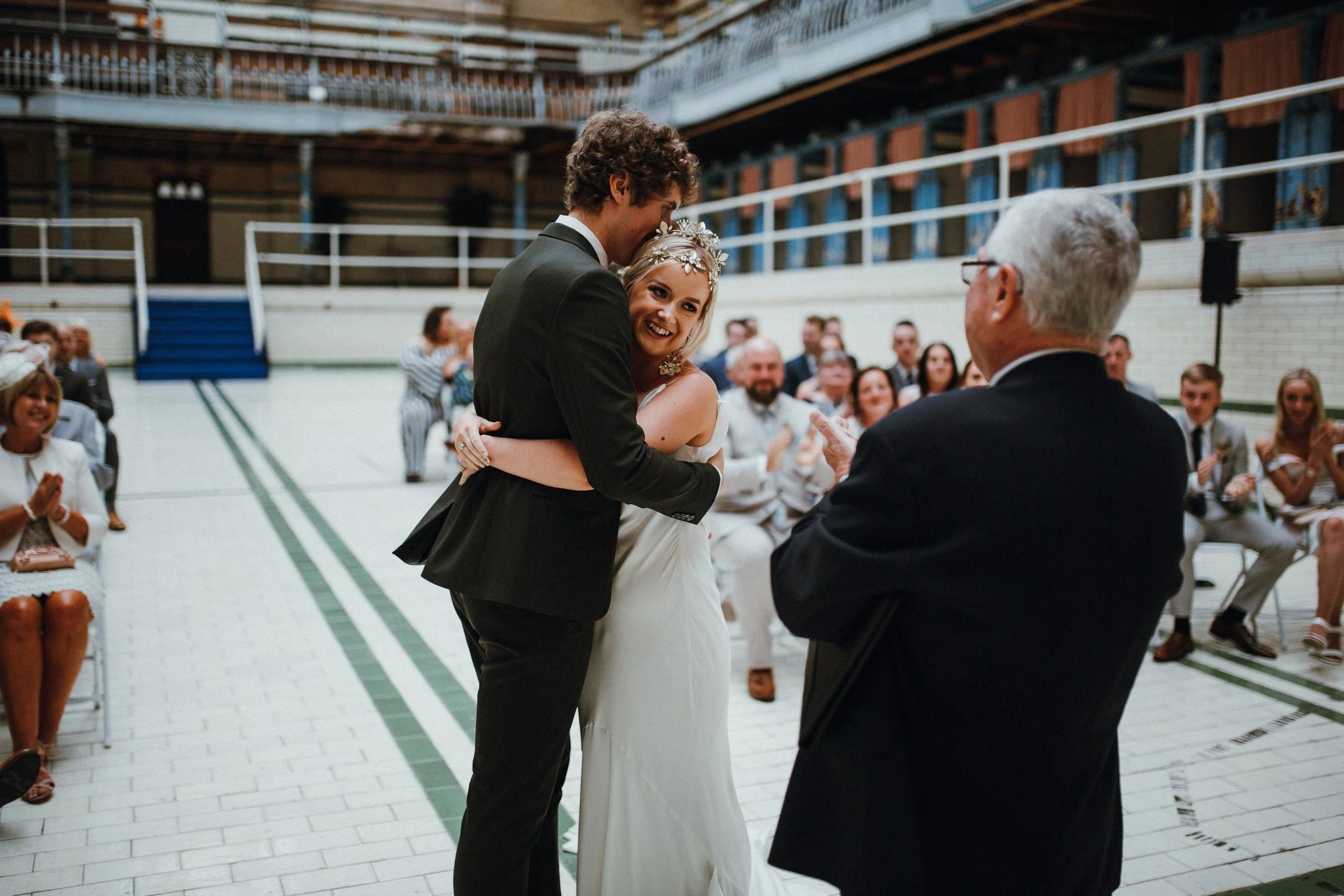 A bride and groom embrace during their wedding ceremony in a large indoor venue, with guests seated and applauding. The bride wears a white dress and a headpiece, while the groom is in a black suit. The setting features tiled floors and ornate railings.