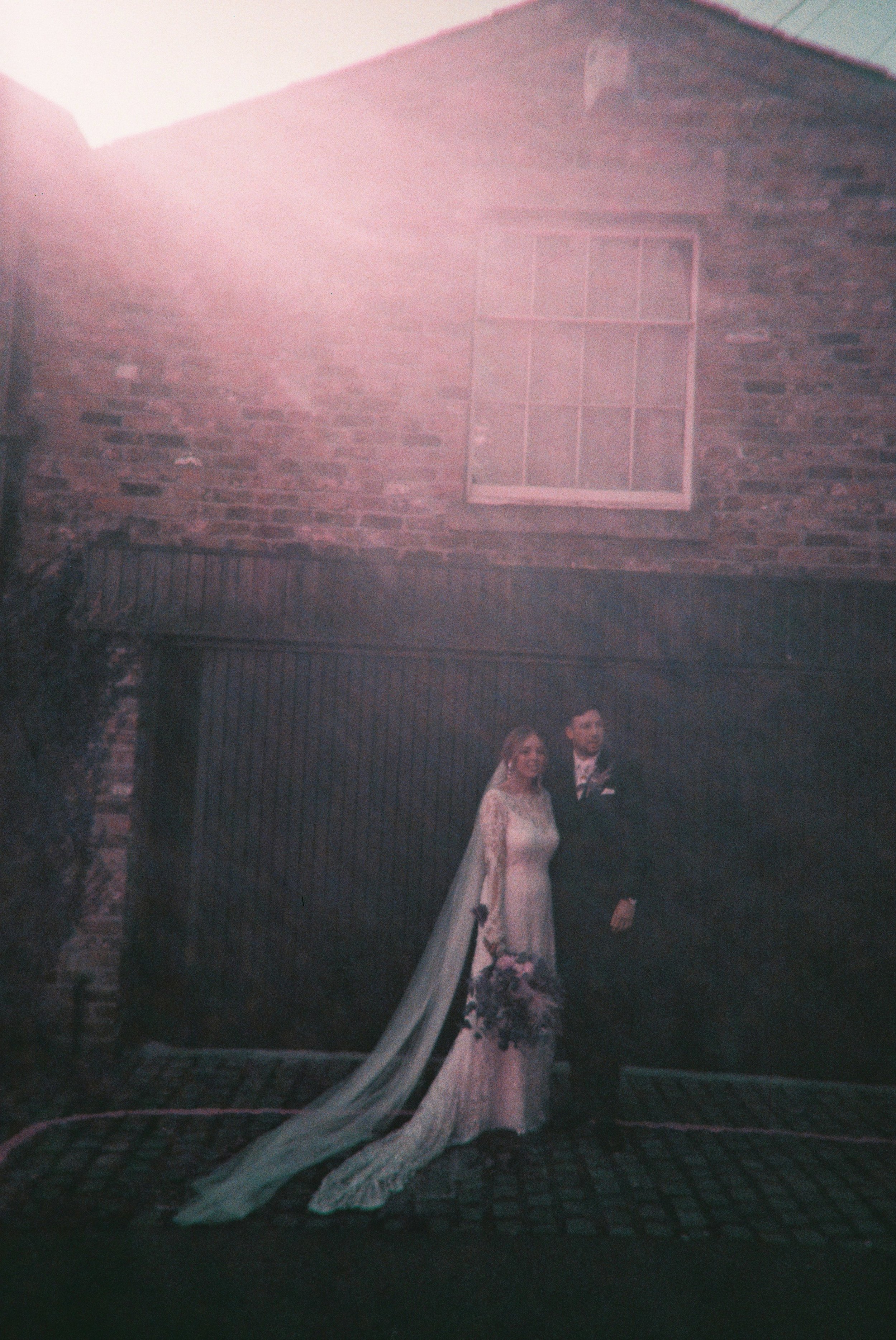 Bride and groom posing in front of a brick building with a large window; bride in a white dress holding a bouquet, groom in a suit, with a shining light effect above them.