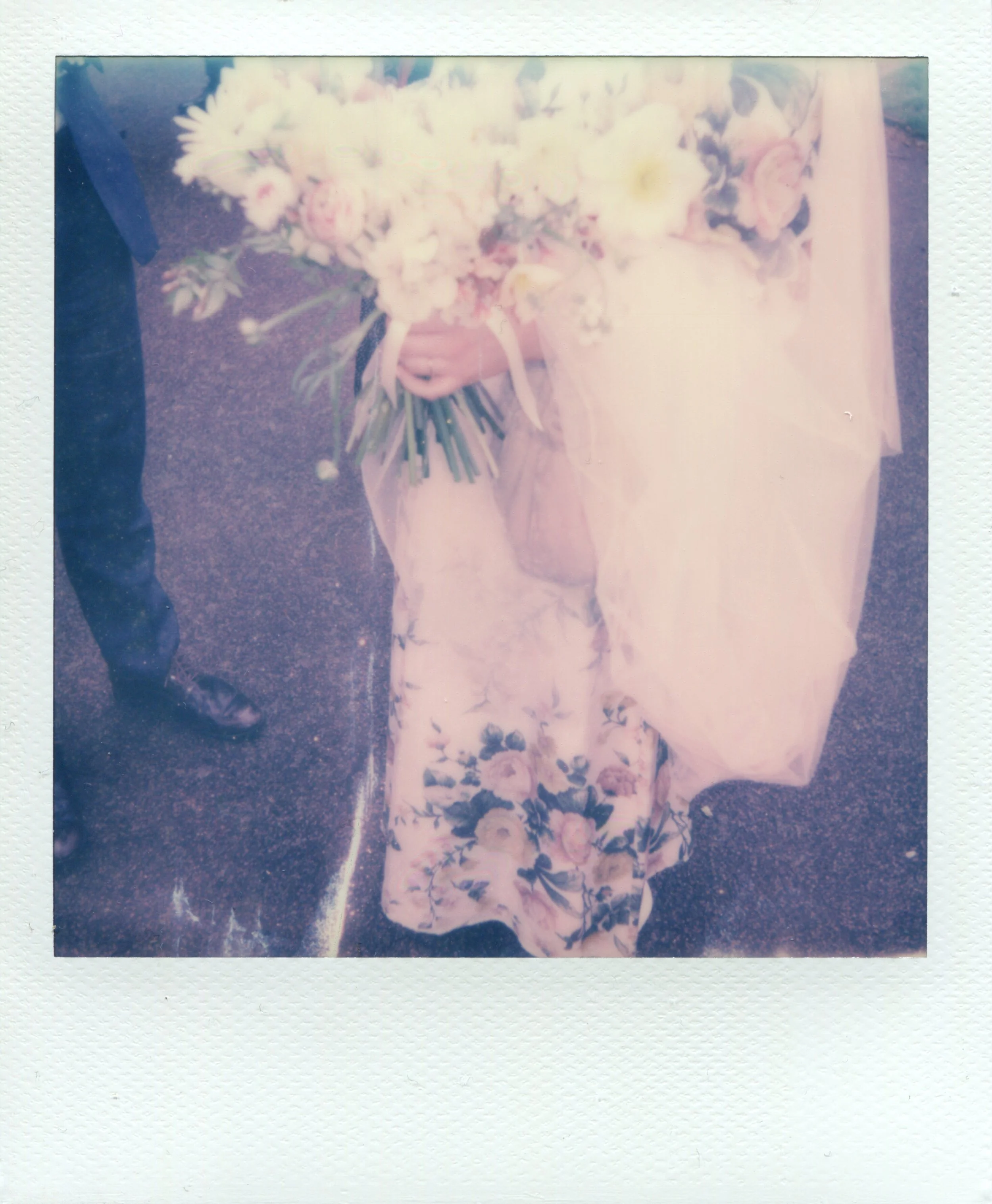 Person in floral dress holding bouquet of flowers, standing on pavement, vintage style Polaroid.