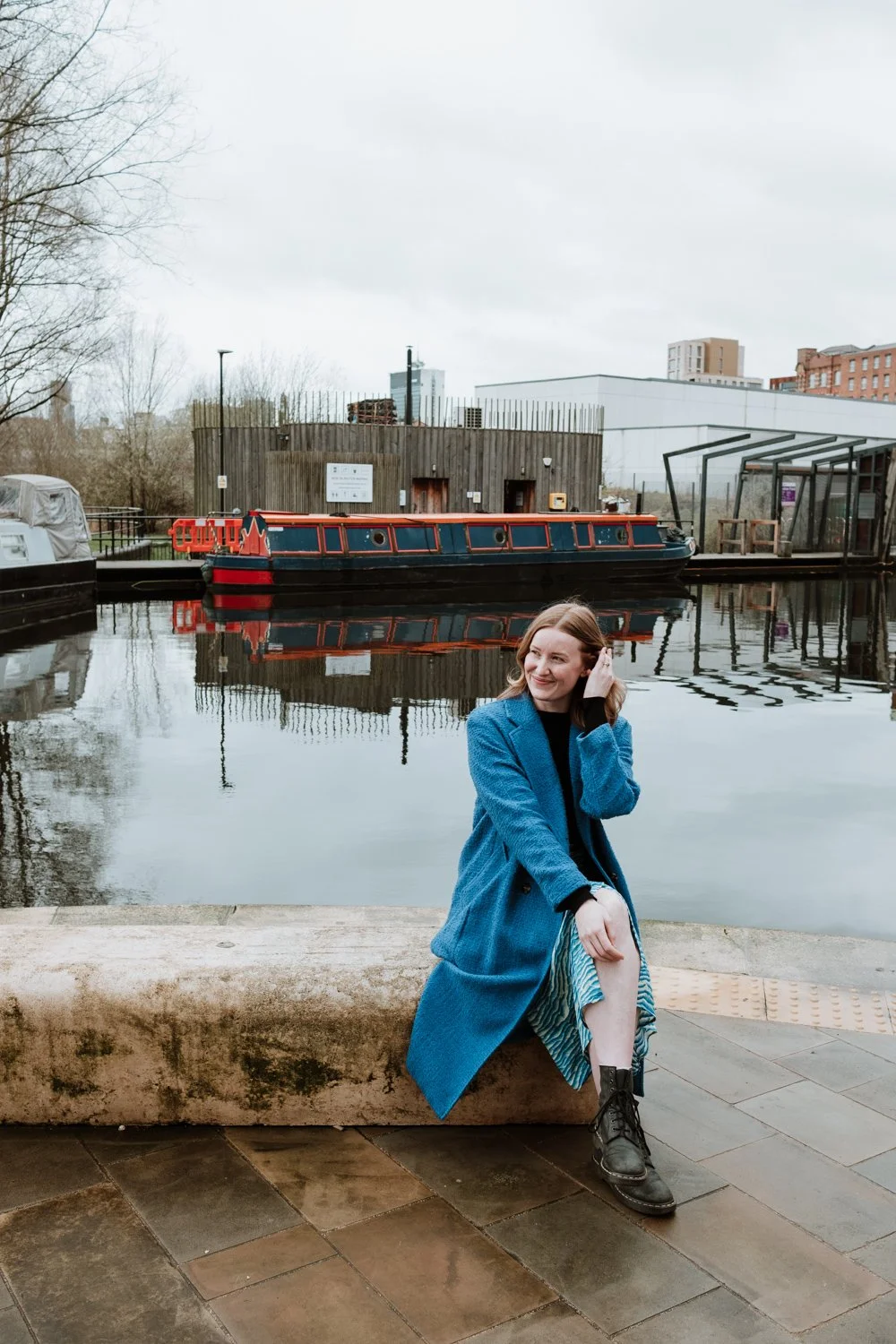 A woman in a blue coat sits by a canal with a narrowboat in the background.