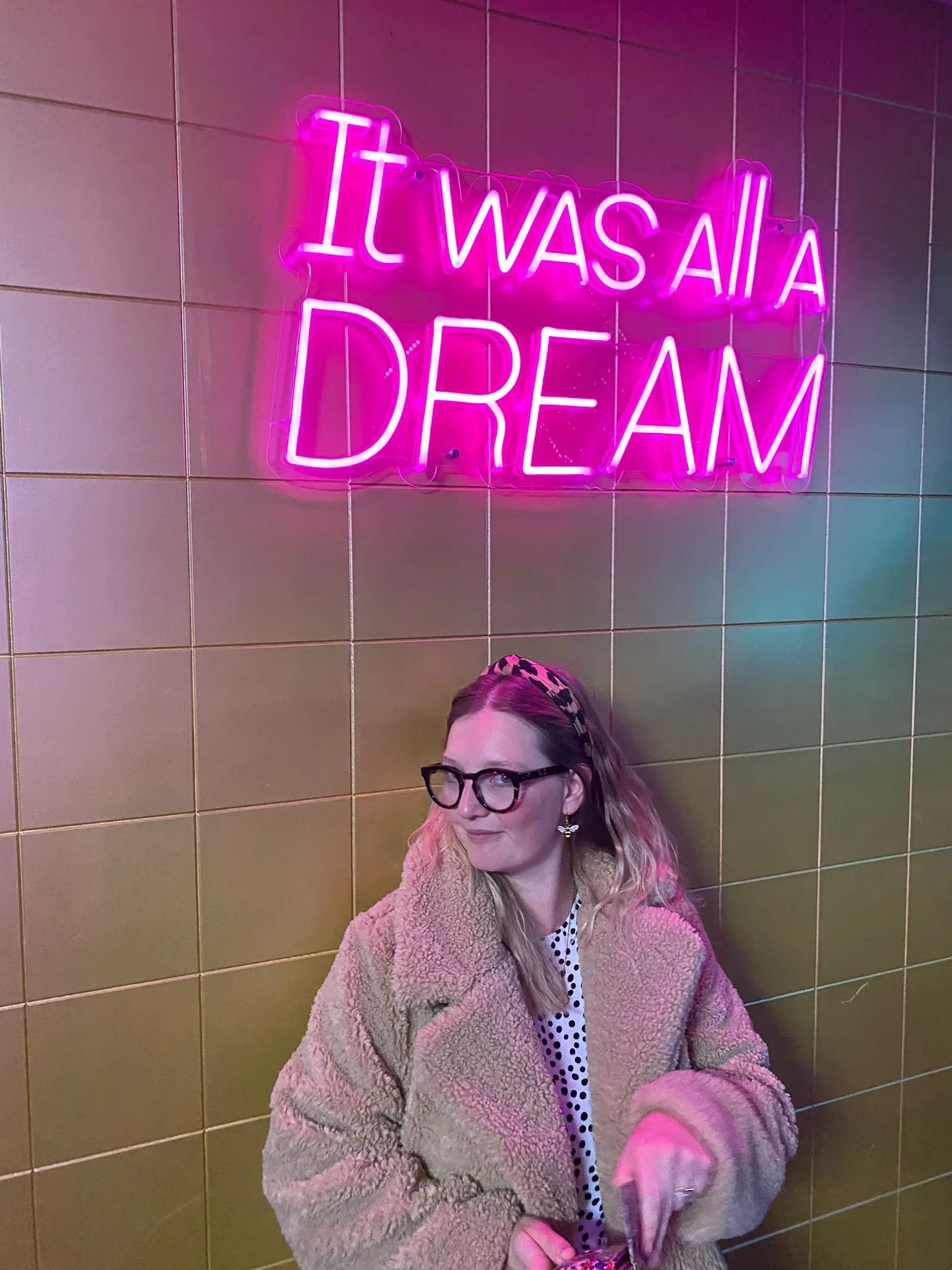 A woman with glasses, wearing a beige furry coat and a polka dot shirt, sitting in front of a yellow tiled wall with a pink neon sign that reads 'It was all a DREAM'.