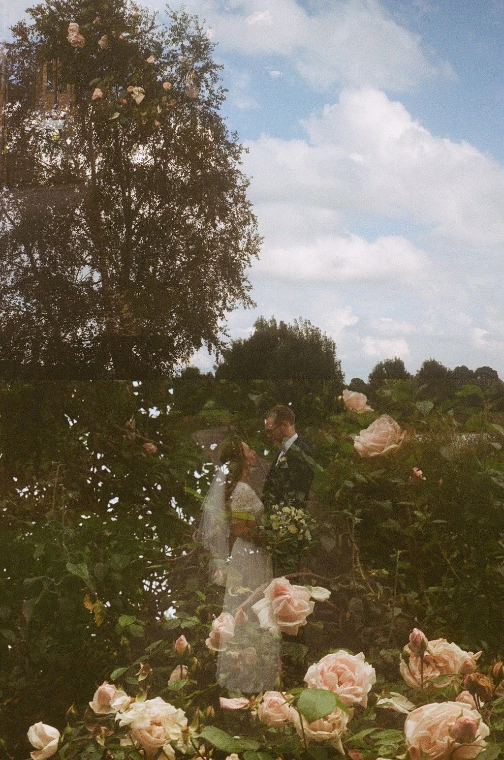 Double exposure photo of a couple in wedding attire among trees and roses with clouds in the background.