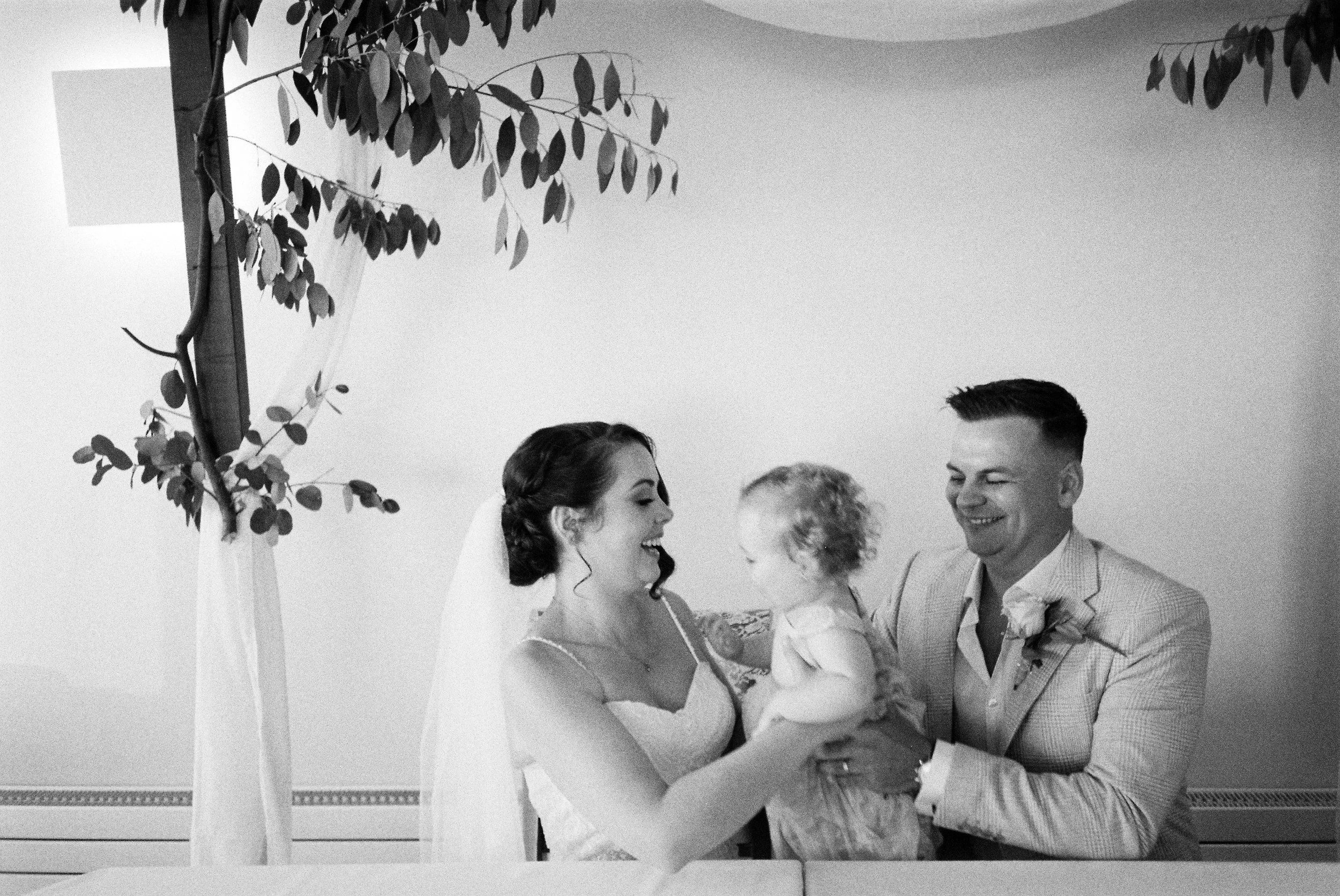 A bride and groom sitting indoors, smiling at a young child they are holding between them. The bride is wearing a veil and a lace dress, while the groom is in a light-colored suit with a boutonniere. There is foliage decor in the background.