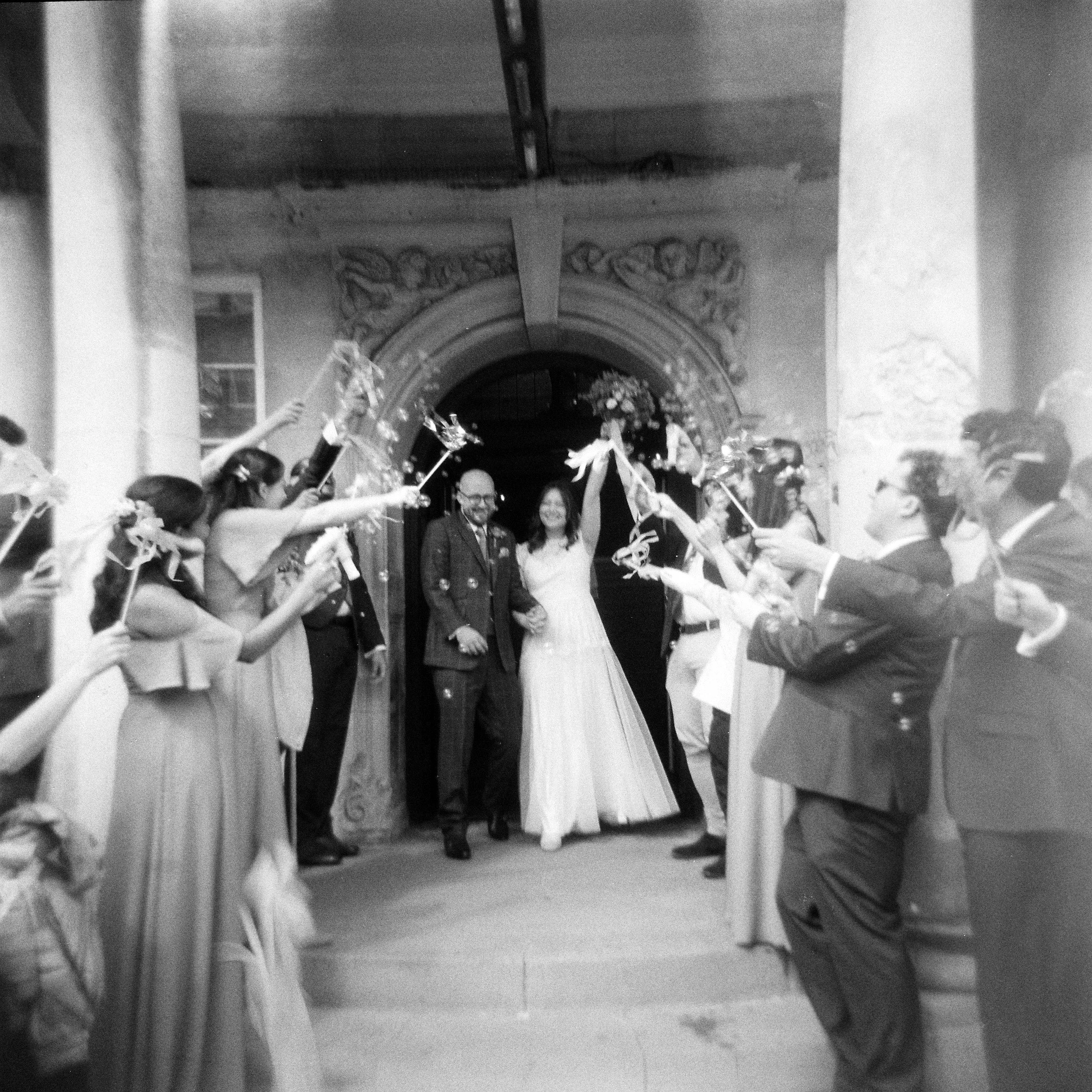 A black and white photo of a couple leaving the Battersea arts centre. Instead of confetti they have bubbles and streamers in the air being held by the wedding party around them