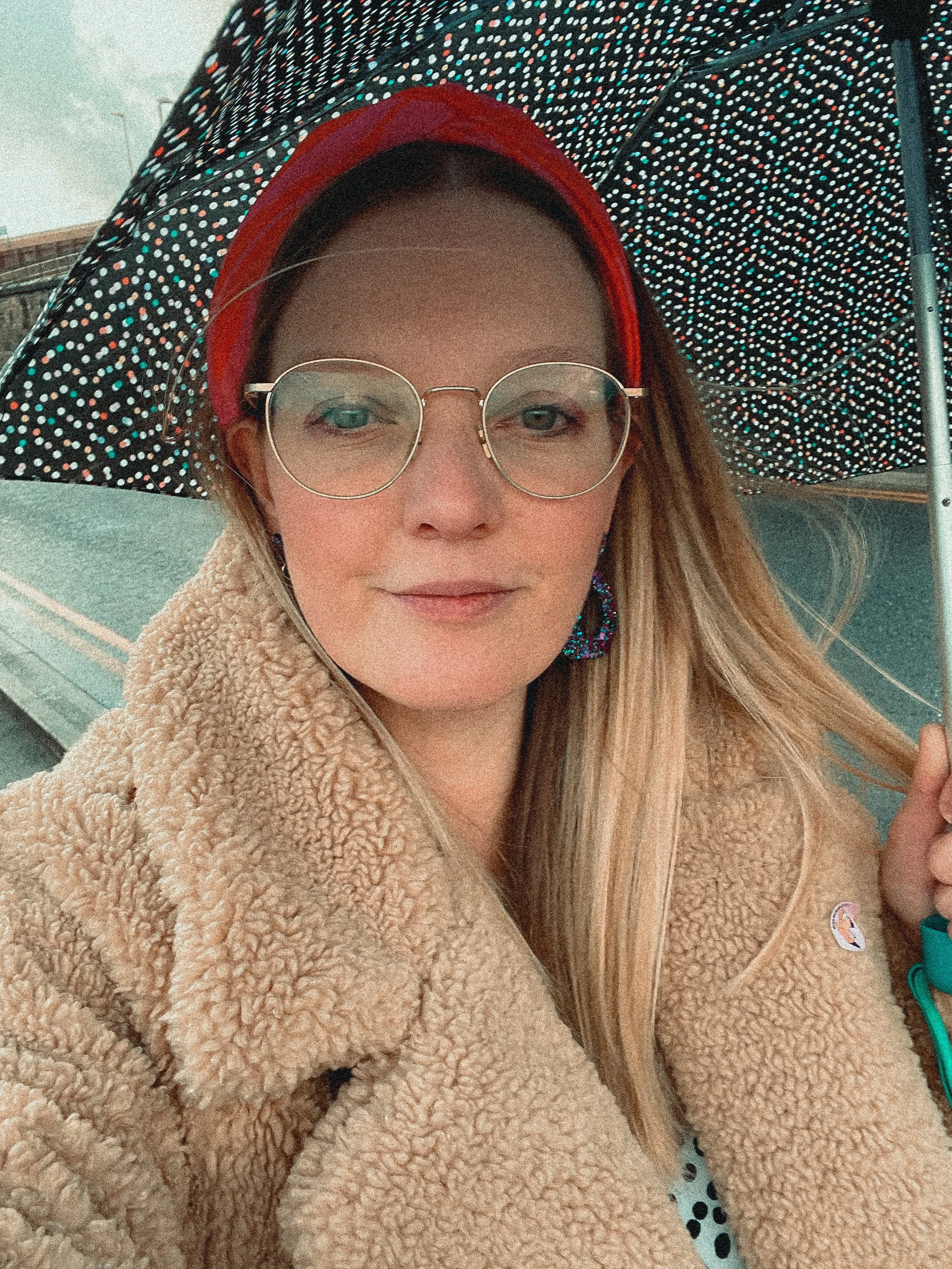 Me (Jenn Davy-Day Photography) looking into the camera, taking a selfie. Wears a fluffy teddy coat, gold round rimmed glasses and has a red headband in her hair. I'm holding a spotty umbrella