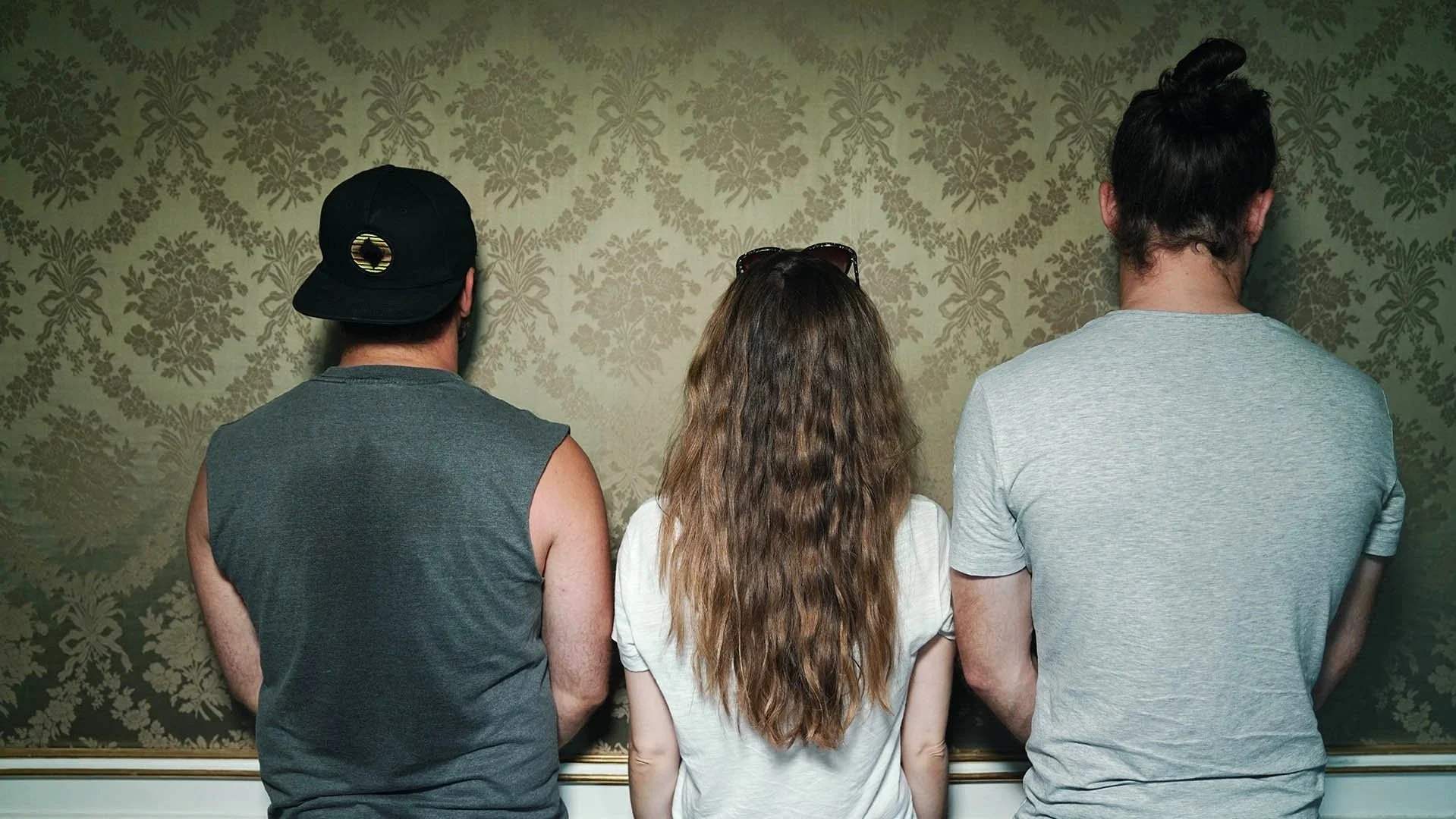 Three people standing with their backs to the camera, looking at a decorative wallpapered wall, two men and a woman, woman has long wavy hair and is wearing sunglasses on her head, man on left wears a cap, man on right has long hair tied back.