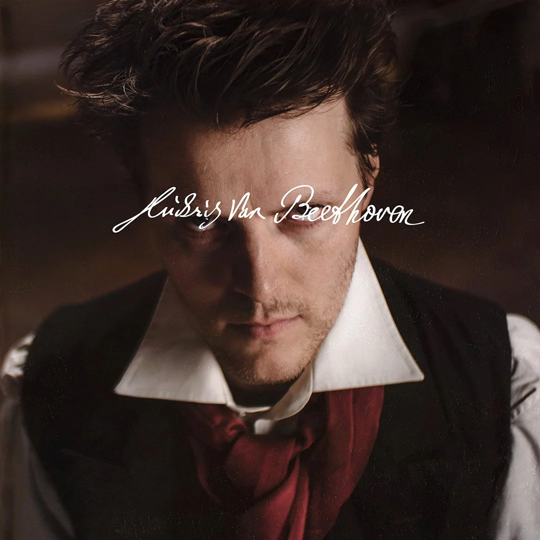 Close-up of a man with styled dark hair, wearing a white shirt, black vest, and red cravat, with a dark background, and the handwritten text 'Ludwig Van Beethoven' across the image.