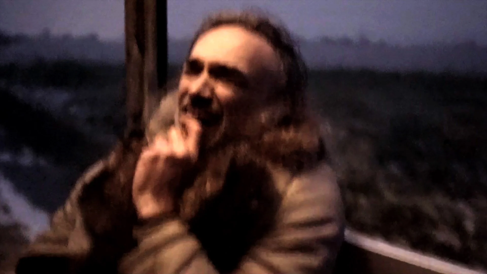 A woman with long curly hair is outside at night, smiling and laughing, with her hand near her face. The background is dark with a faint landscape or field.