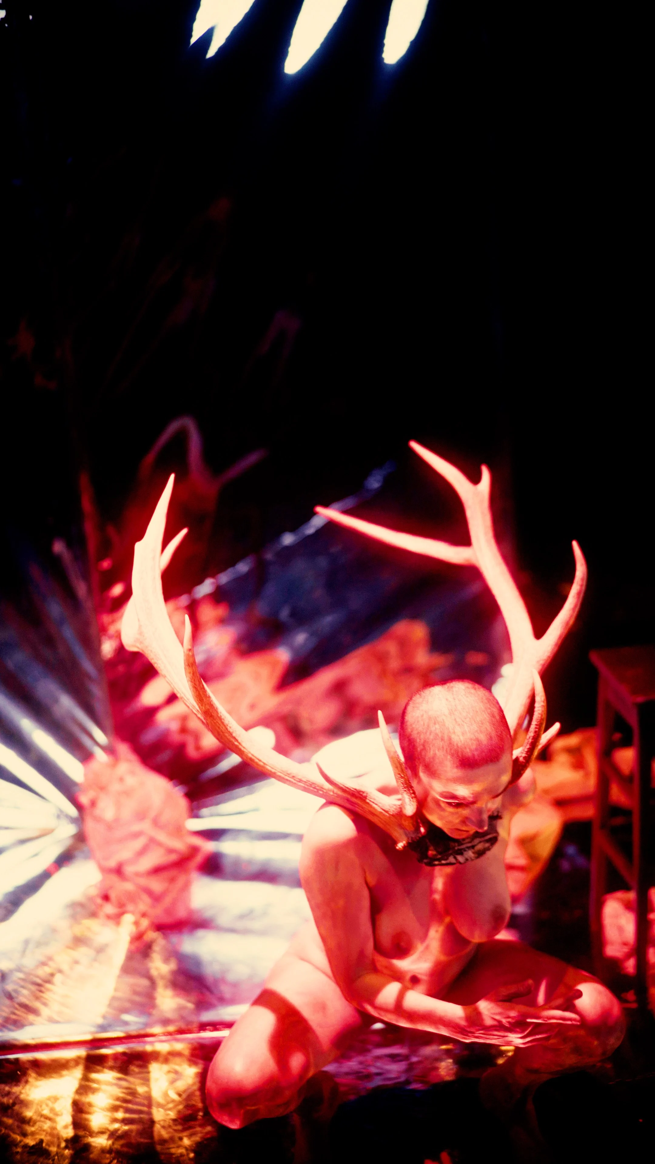 A person with a painted face, wearing antlers, kneeling on the ground in front of a Halloween or fantasy-themed display with bright, colorful lights and decorations.