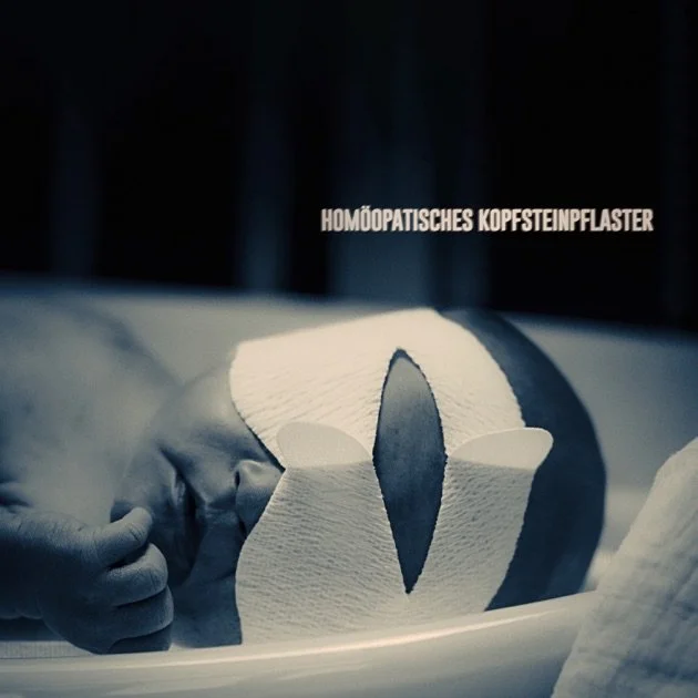 A newborn baby with a homoeopathic head cap plaster on their head, lying on a medical table or bed.