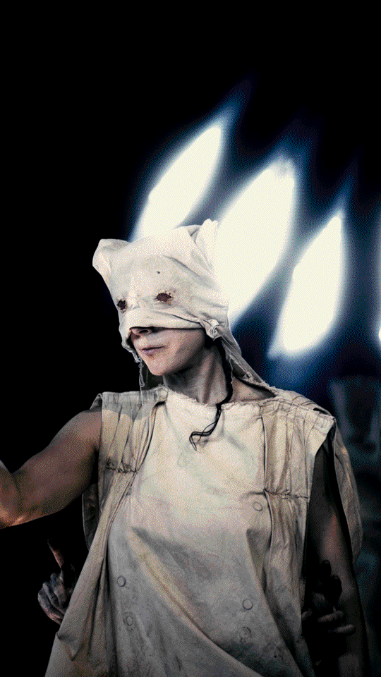 A person wearing a pale costume resembling a mummy, with a cloth covering their eyes, standing in front of bright, blurred lights.
