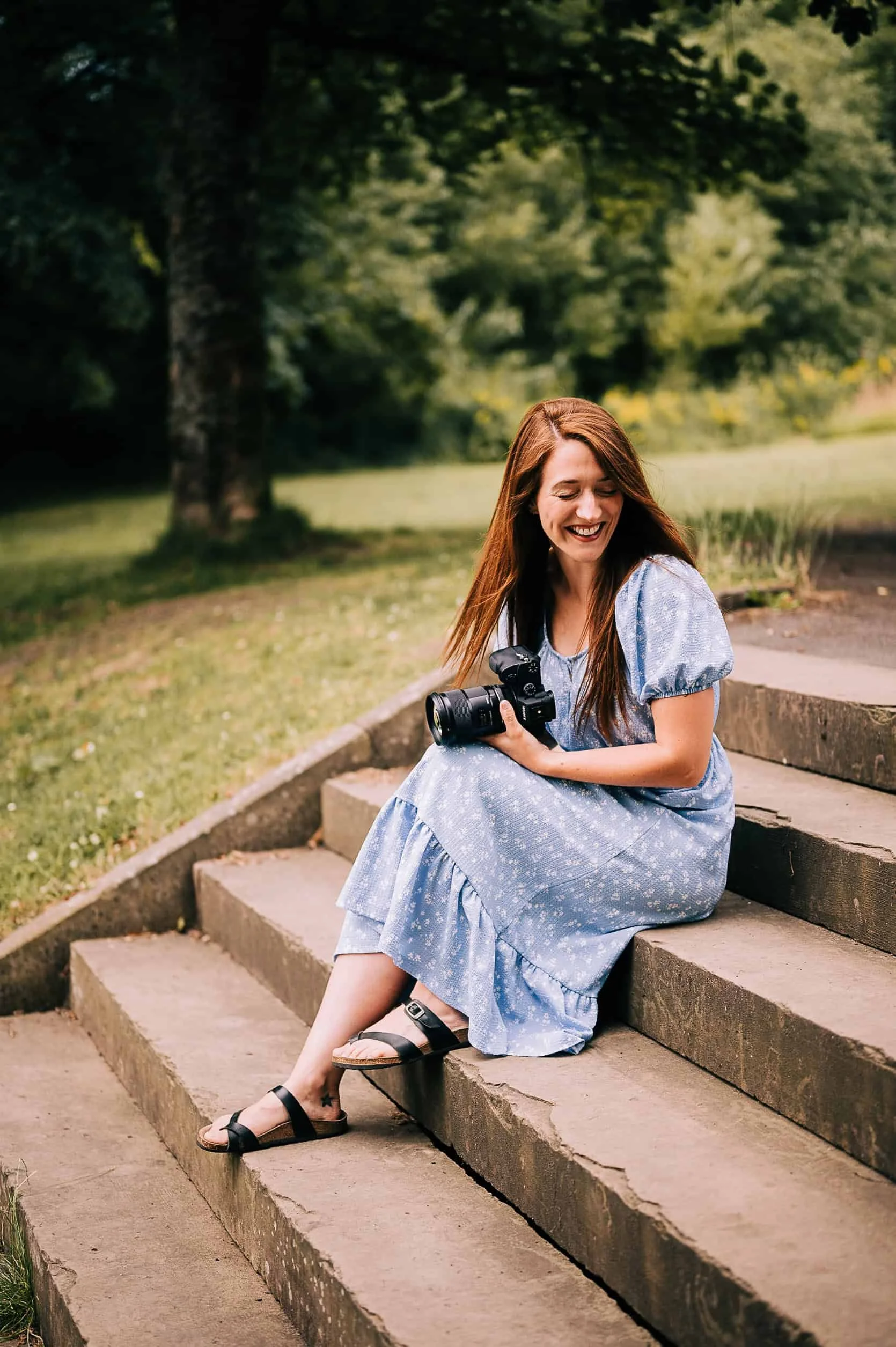 Clare robinson photography sat in a set of stairs in a park holding camera and laughing