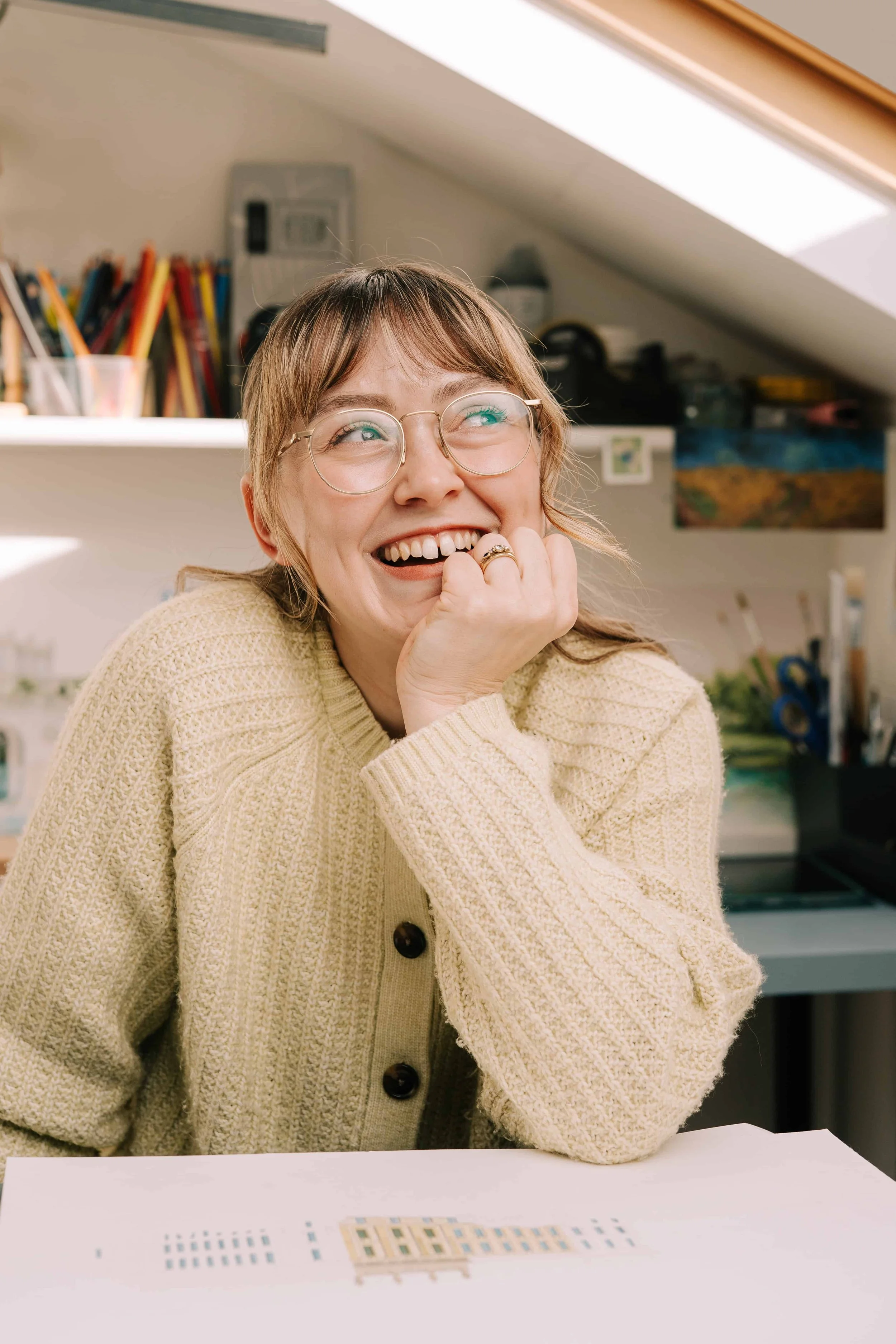 Fine artist lydia sat in her home studio with a work in progress drawing of Chatsworth in front of her and he work area behind her