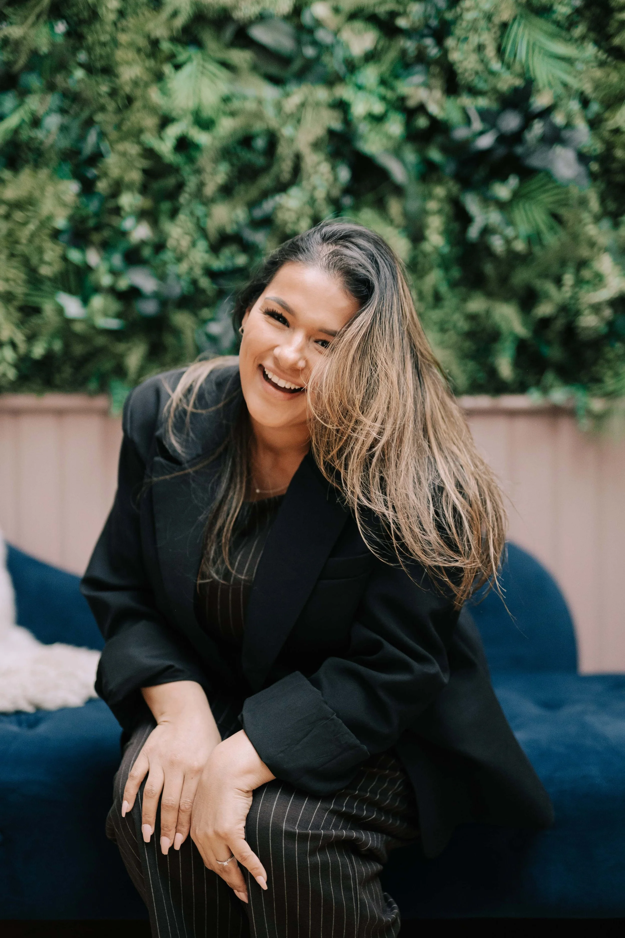 Content creator in smart black outfit sat on velvet blue sofa with her hair to one side and a beaming smile