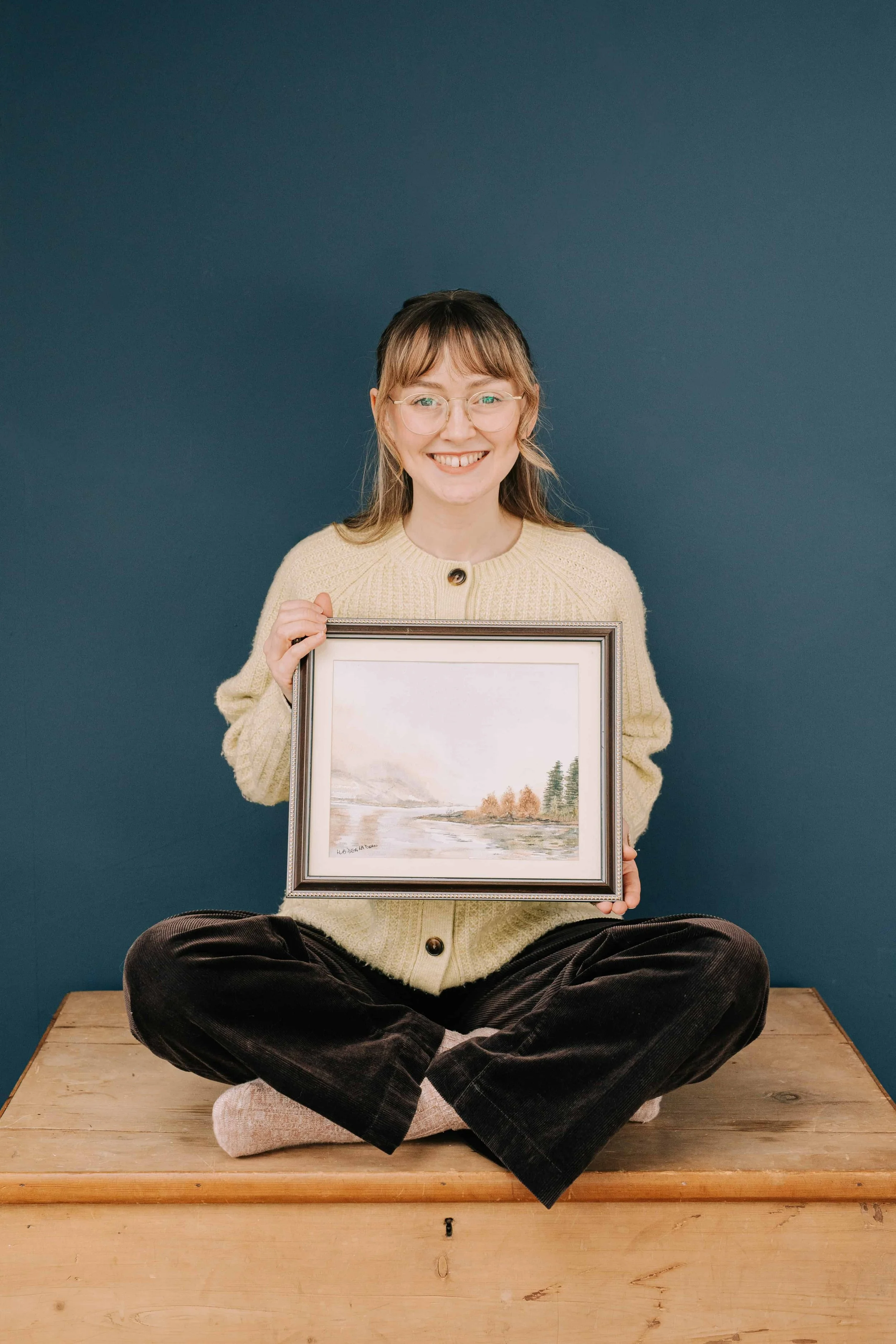 Lydia Draws sat on wooden box holding her favourite painting by her grandfather