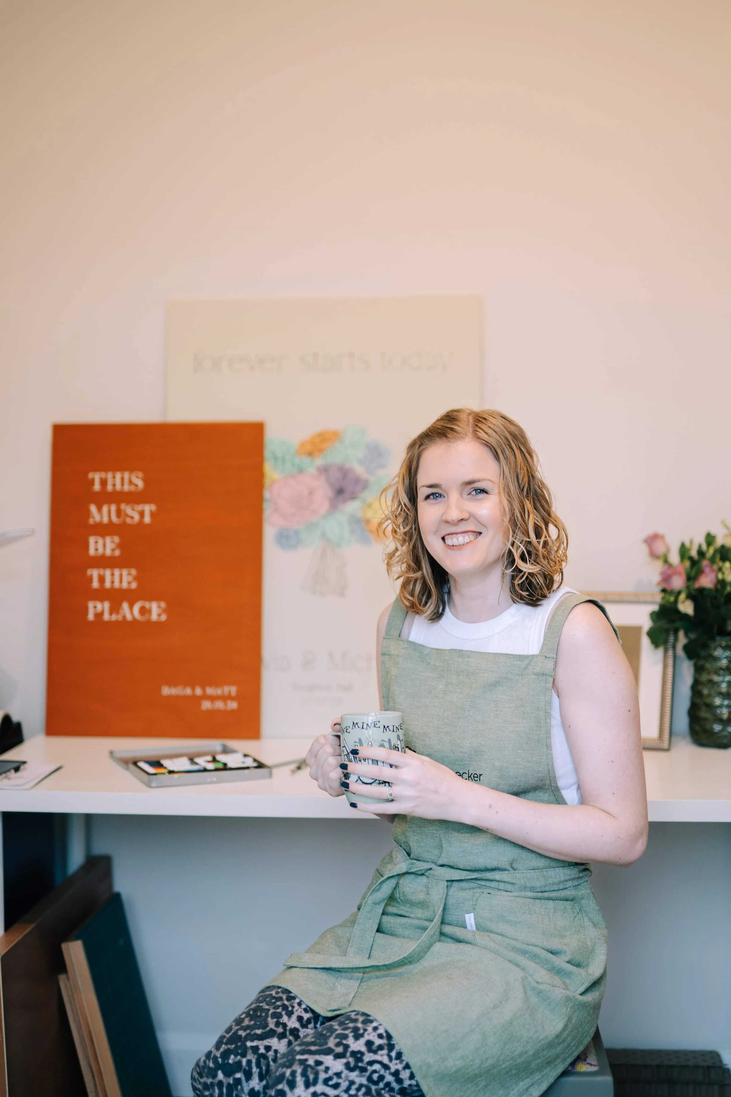 Woodpecker by designs sat in her painting studio with green apron, holding a cup of tea