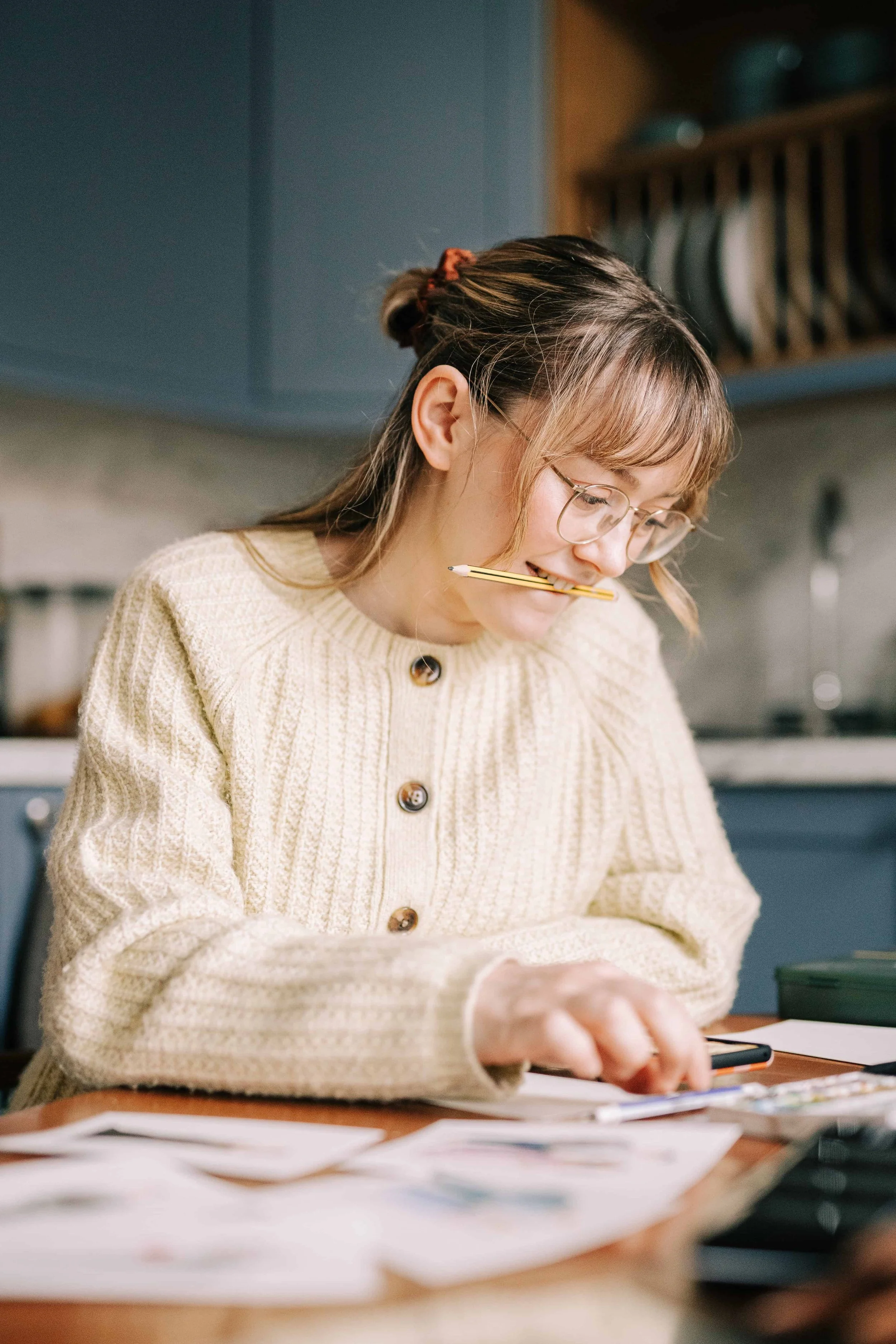 Lydia Draws sat at table painting with a coloured pencil in her mouth