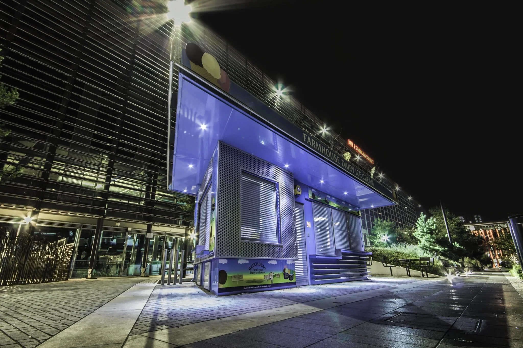 Nighttime shot of an illuminated blue Dairy Ice Cream kiosk with stairs leading up to the entrance, set against a modern building with metal and glass facade, trees, benches, and streetlights. photography by shanna may in Birmingham