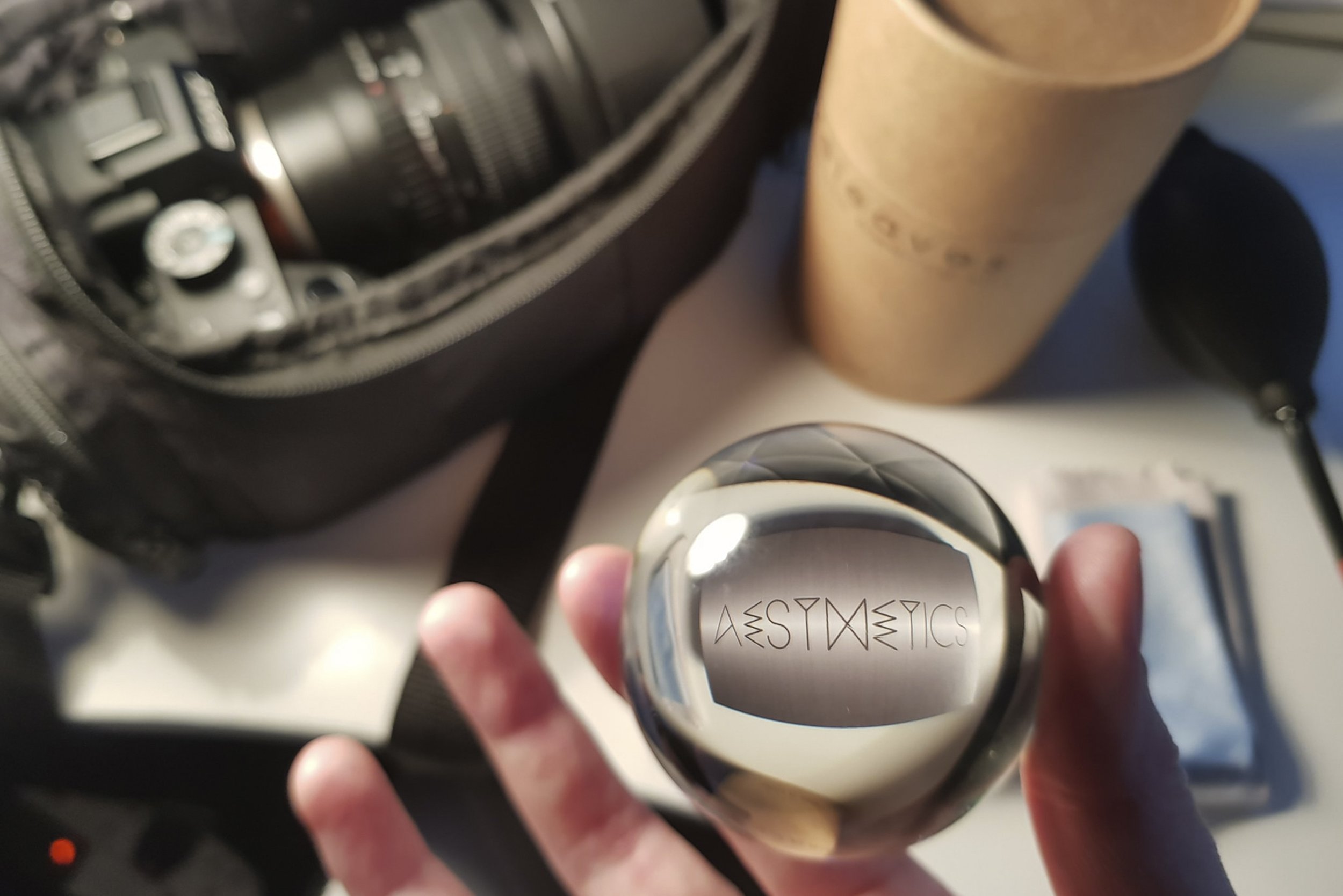 A person holding a reflective, metallic perfume bottle with the brand name 'Aesthetics' engraved on the bottom. The background includes a black bag, a brown container, and various items on a white surface.