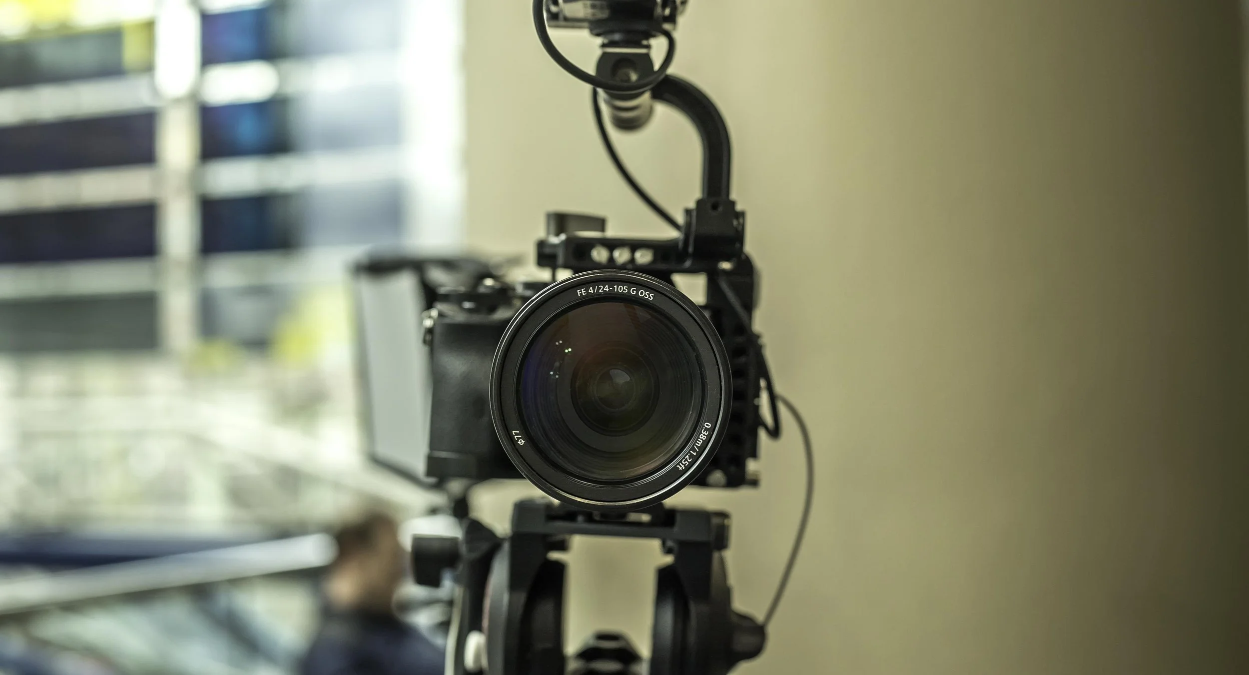 Close-up of a professional camera mounted on a tripod, with a blurred background including a person and a grid-like structure. photography by shanna may in new york city