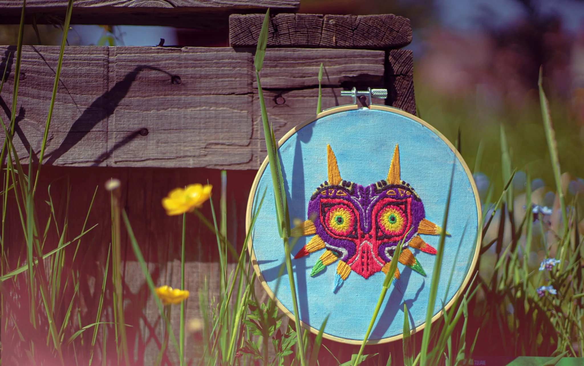 Embroidery hoop with a colorful butterfly design resting against a weathered wooden fence, surrounded by tall green grass and small yellow flowers. photography by shanna may