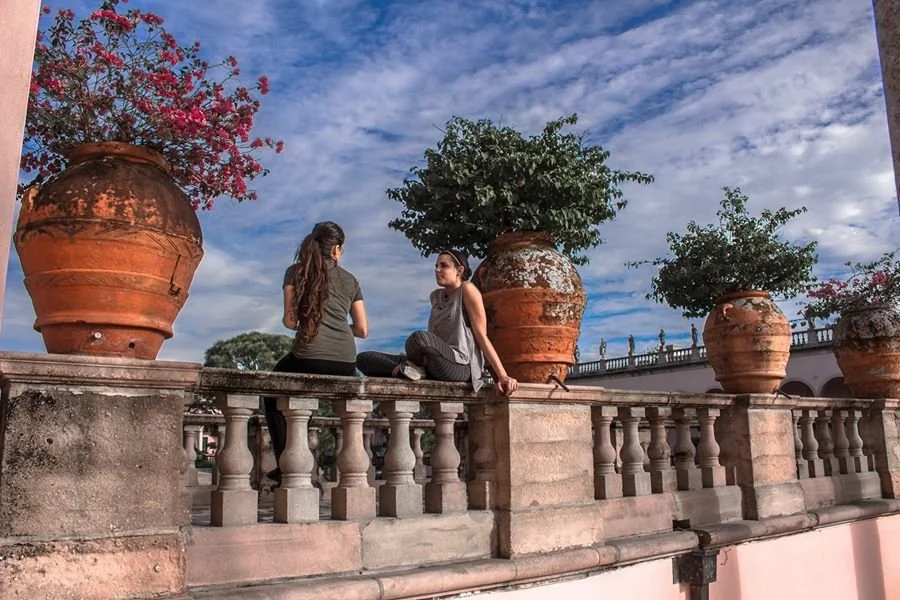 Two women sit on a stone railing with large clay pots and trees behind them, under a sky with clouds photography by shanna may in Florida
