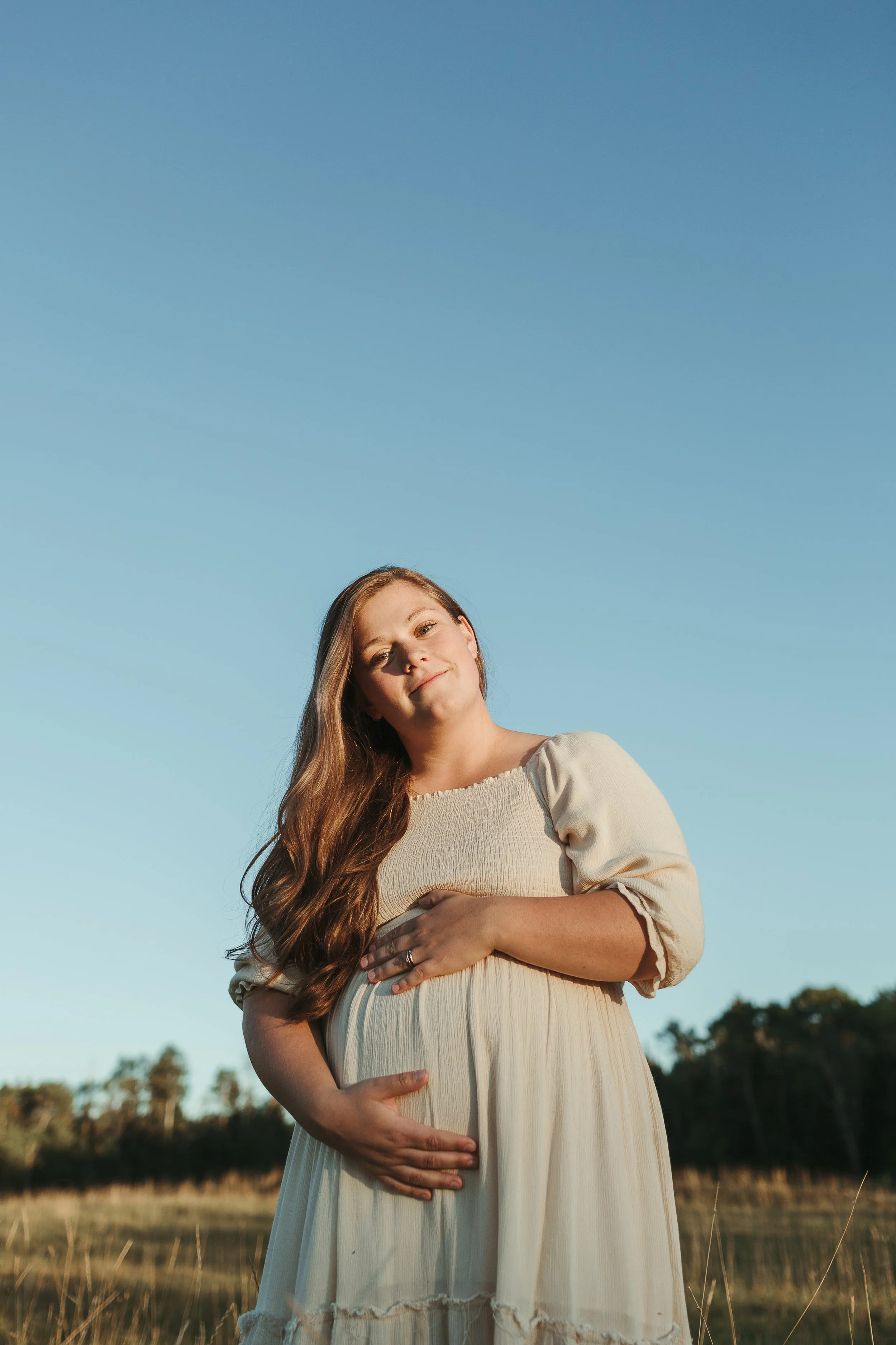 A pregnant woman with long brown hair standing outdoors in a field under a clear blue sky, gently holding her belly with one hand, during late afternoon or early evening.