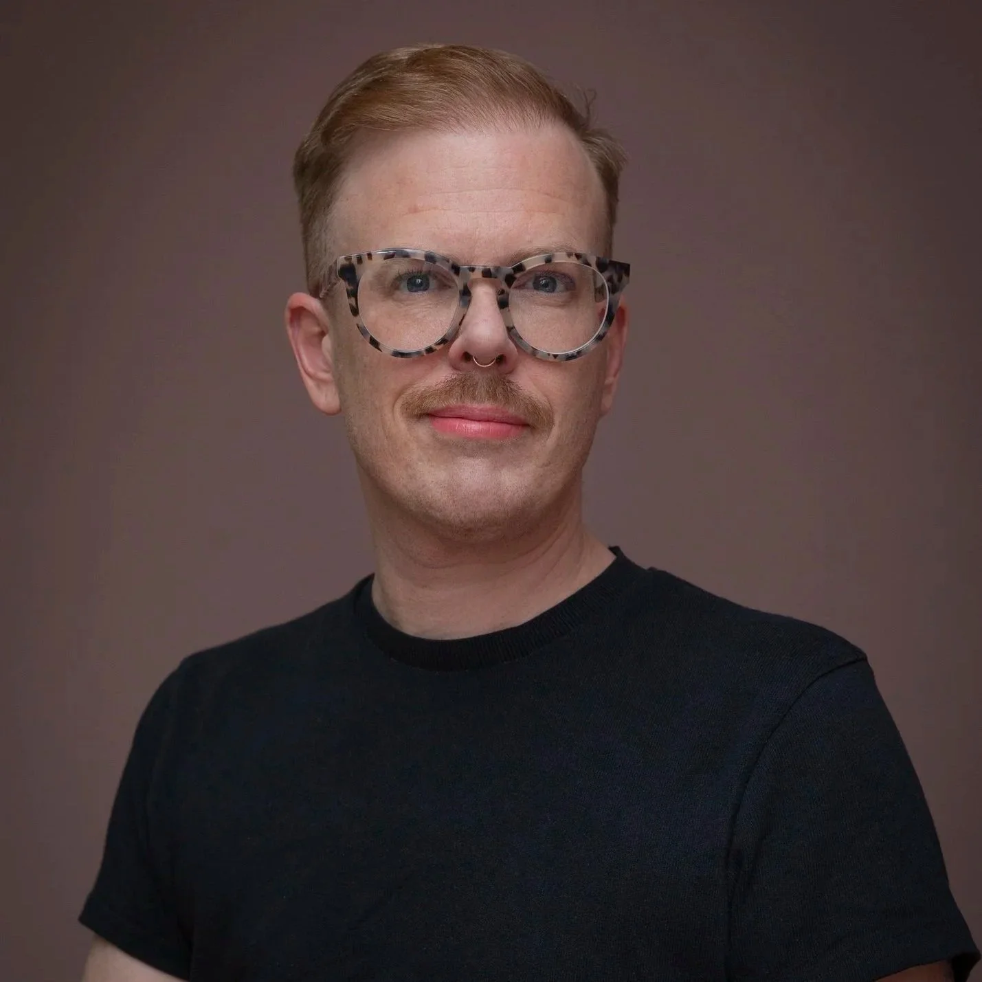 A person with short, reddish hair, wearing large tortoiseshell glasses and a black shirt, smiling slightly against a plain background.