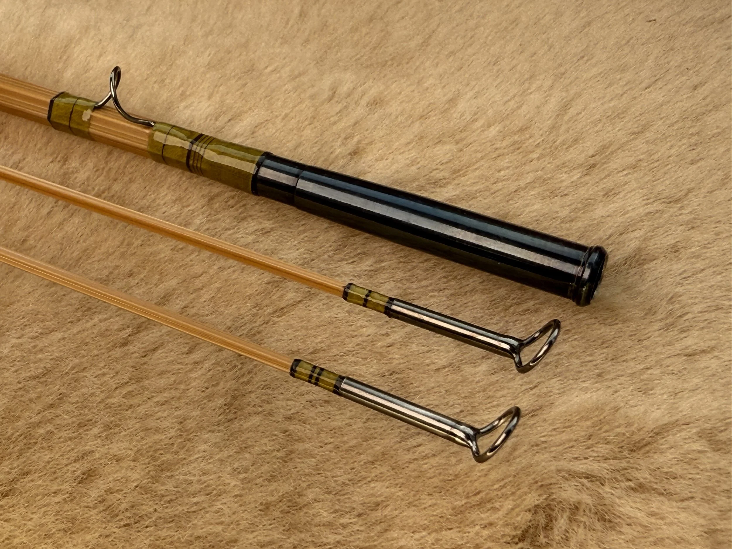 Three blonde bamboo fly rod sections laid on a sheepskin surface, with the butt section showing a blued nickel silver ferrule and a snake guide with green thread wraps and single turn brown inlays, and the two tip sections displaying dark nickel tip-