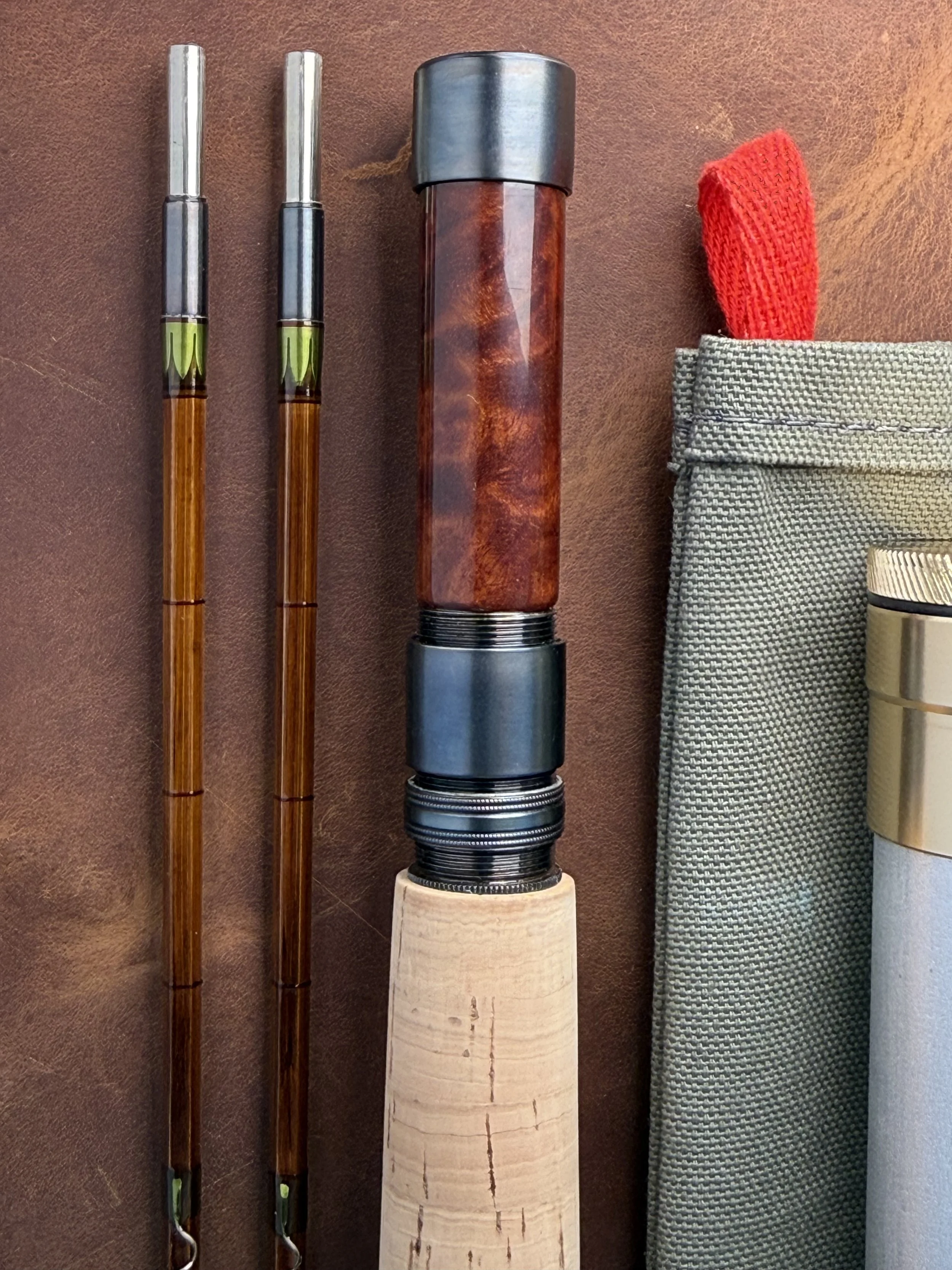Bamboo rod close up featuring reel seat, part of cork grip, two tip sections, rod bag with red pull and edge of rod tube.