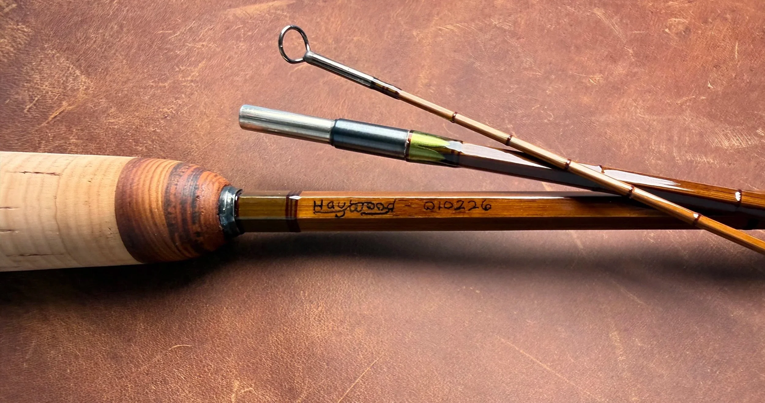 Close-up of a handcrafted bamboo fly rod handle area showing a cork grip, cottonwood ring at the front, and the maker's inscription "Haywood - 010226" written on the blank, with the tip sections,  snake guide and ferrule visible in the background.