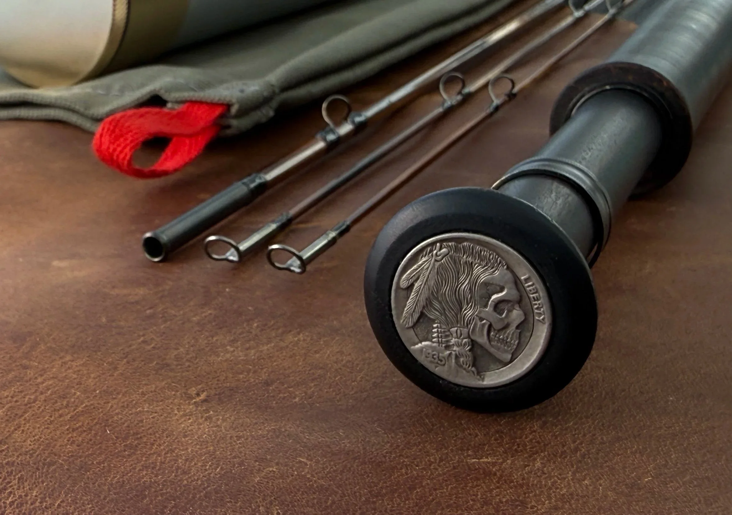 A heavily flamed bamboo fly rod featuring a distinctive butt cap inlaid with a hobo nickel — a hand-carved Buffalo nickel depicting a skull — displayed in front of a canvas rod bag with a red pull and the mid and twin tip rod sections visible in the 