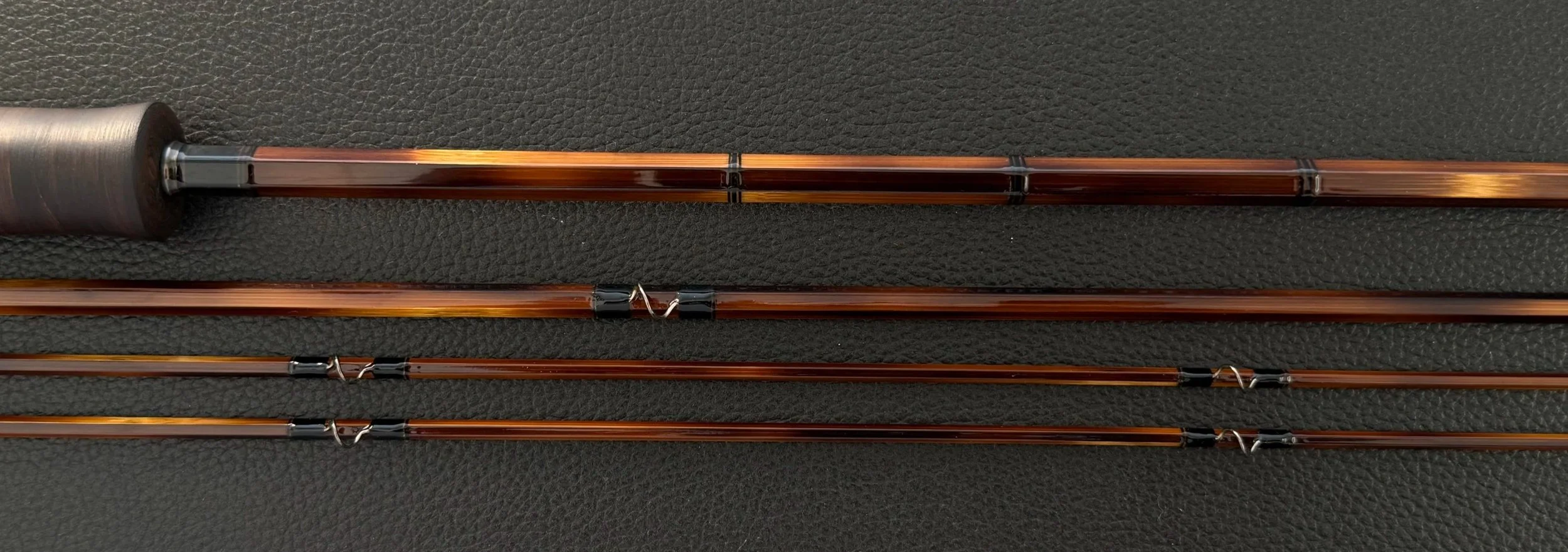Four sections of a handcrafted bamboo fly rod laid horizontally on a dark leather surface, showing the heavily flamed hexagonal blanks with black thread-wrapped snake guides, and a partial view of an ebonized cottonwood grip at the left edge.