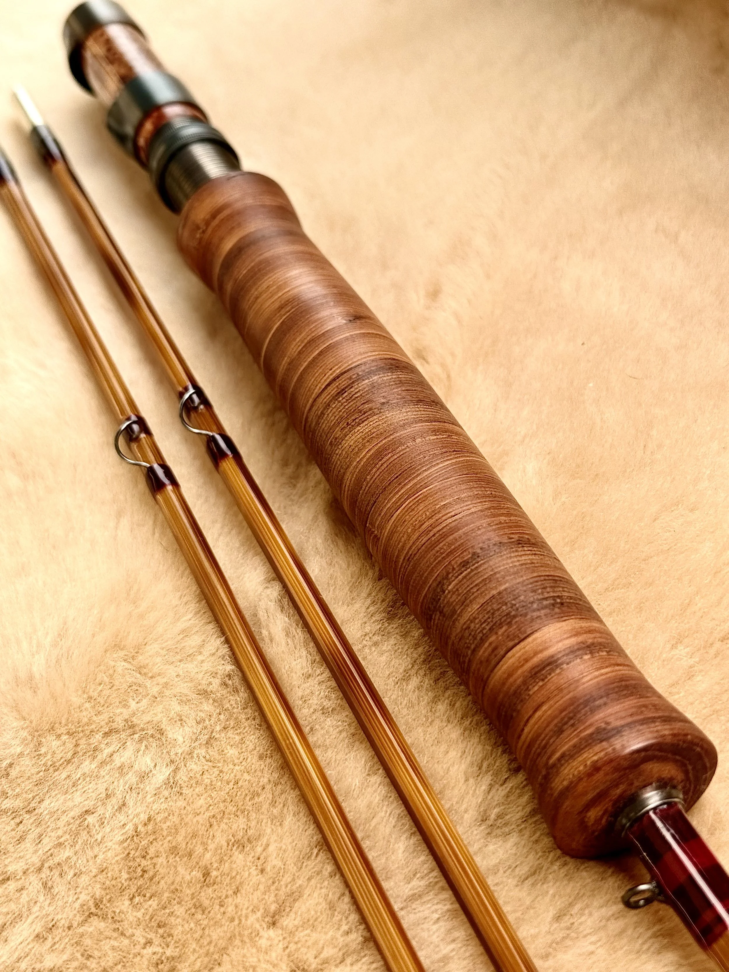 Close-up of a handcrafted bamboo fly rod handle featuring cottonwood bark with warm reddish-brown tones, a figured burl wood reel seat with blued locking hardware, and two flamed tip sections with burgundy thread wraps and snake guides visible below,