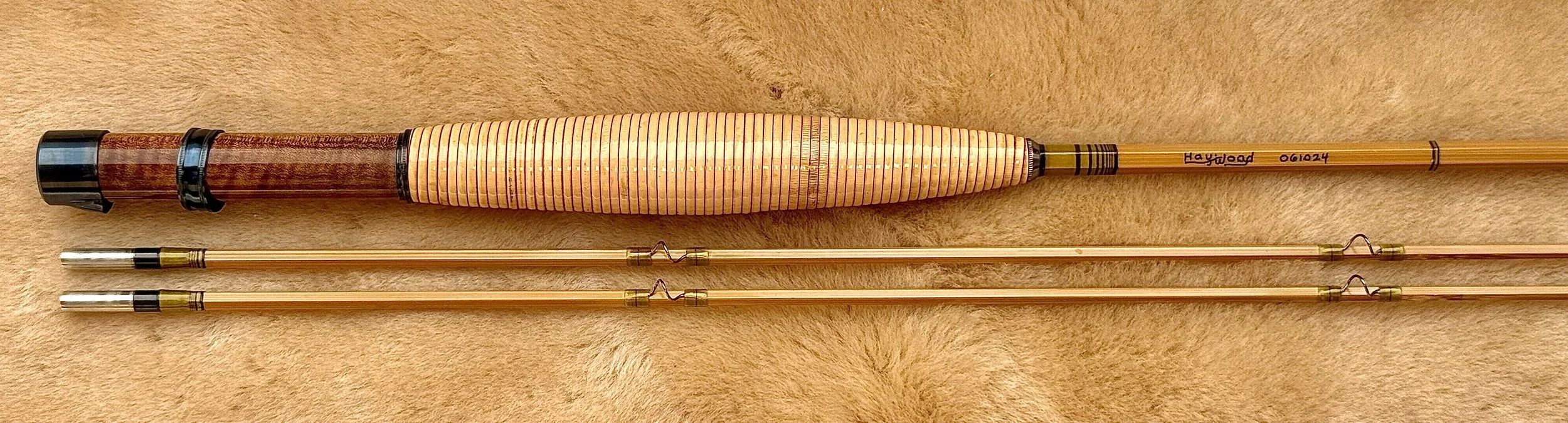 A Haywood bamboo fly rod labeled "Haywood 061024" laid on a light sheepskin surface, featuring a rattan grip, a reddish-brown figured wood reel seat with blued hardware, and two tip sections with nickel silver ferrules, snake guides, and green thread