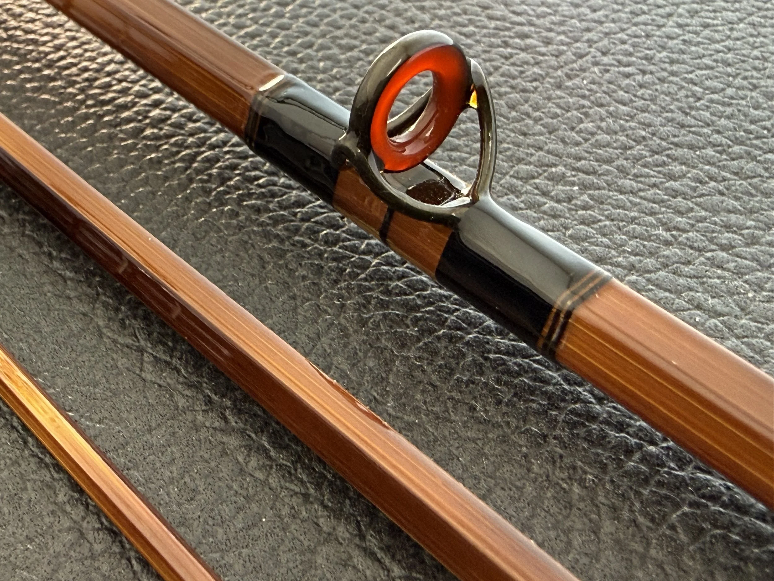 Close-up of a bamboo fly rod tip section featuring a blued stripping guide with a deep red agate ring insert, set against finely varnished hexagonal cane with dark wrappings, alongside two additional rod sections on a dark leather background.