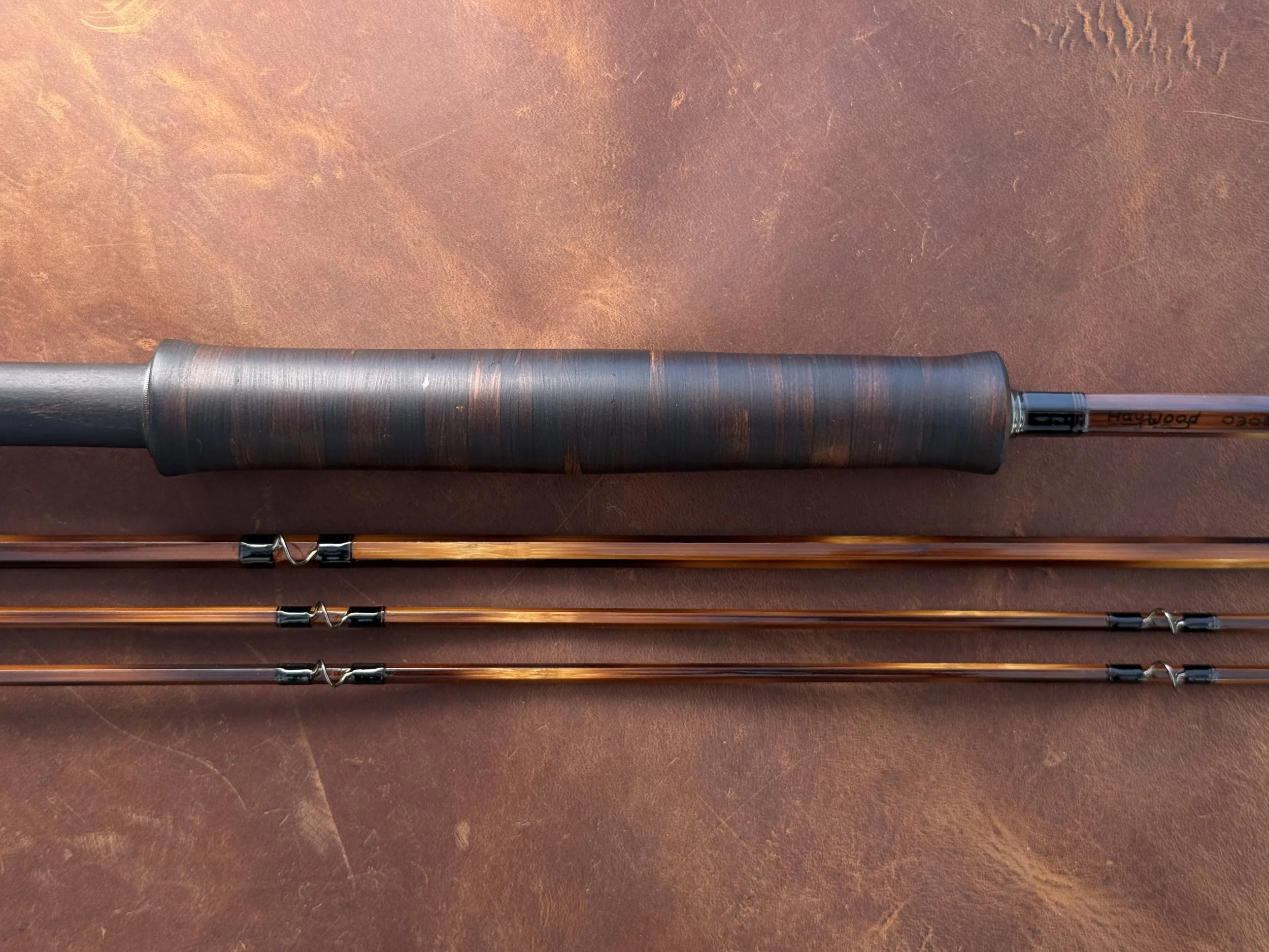 A handcrafted bamboo fly rod grip made ebonzied cottonwood bark with three assembled rod sections below showing amber-toned blanks, black thread wraps, and blued snake guides, with the maker's name "Haywood" visible near the grip.