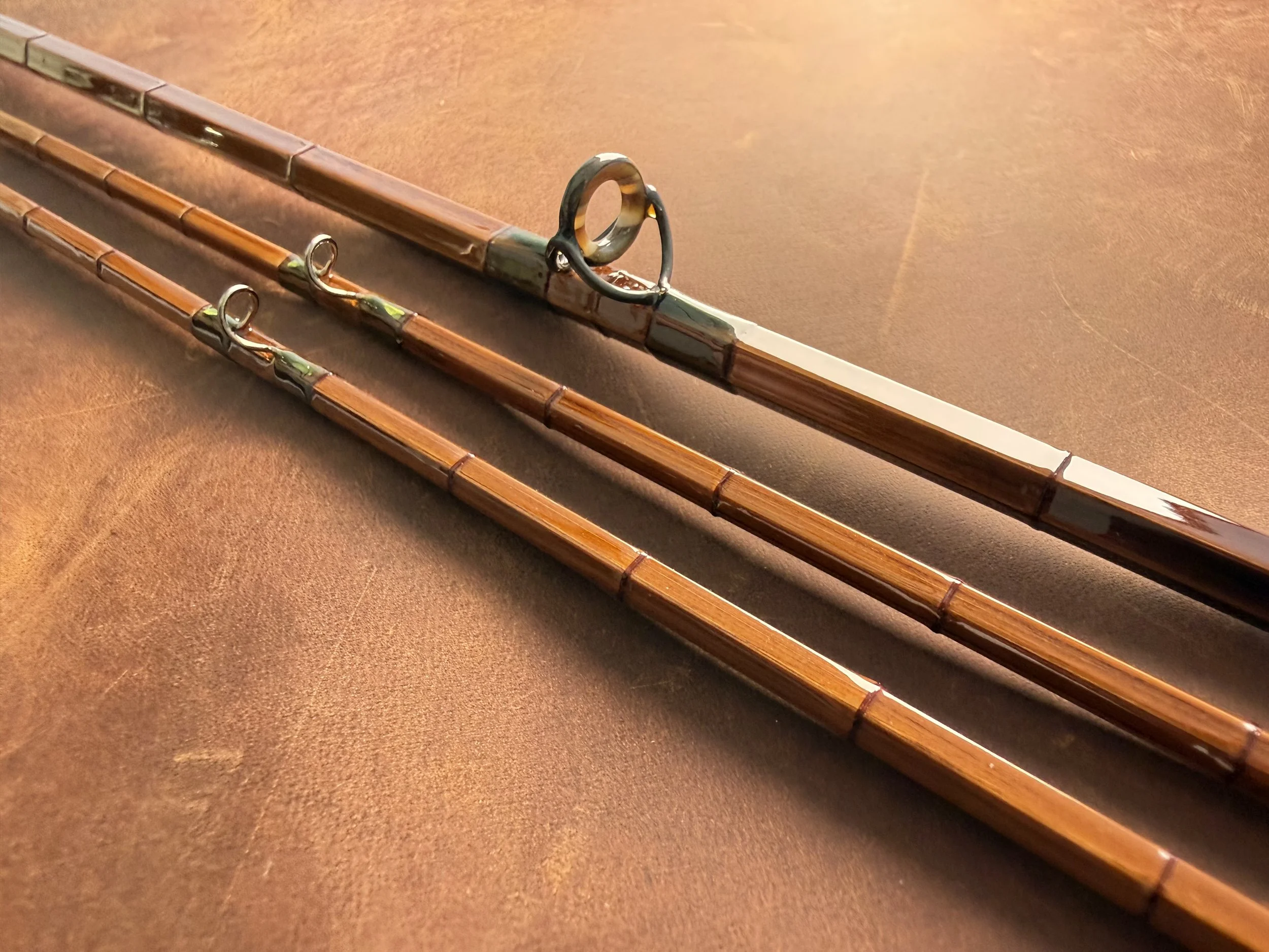 Three sections of a handcrafted bamboo fly rod laid side by side on a leather surface, showing the hexagonal cane construction, olive green and copper wrappings, and blued snake guides, including an agate stripping guide.