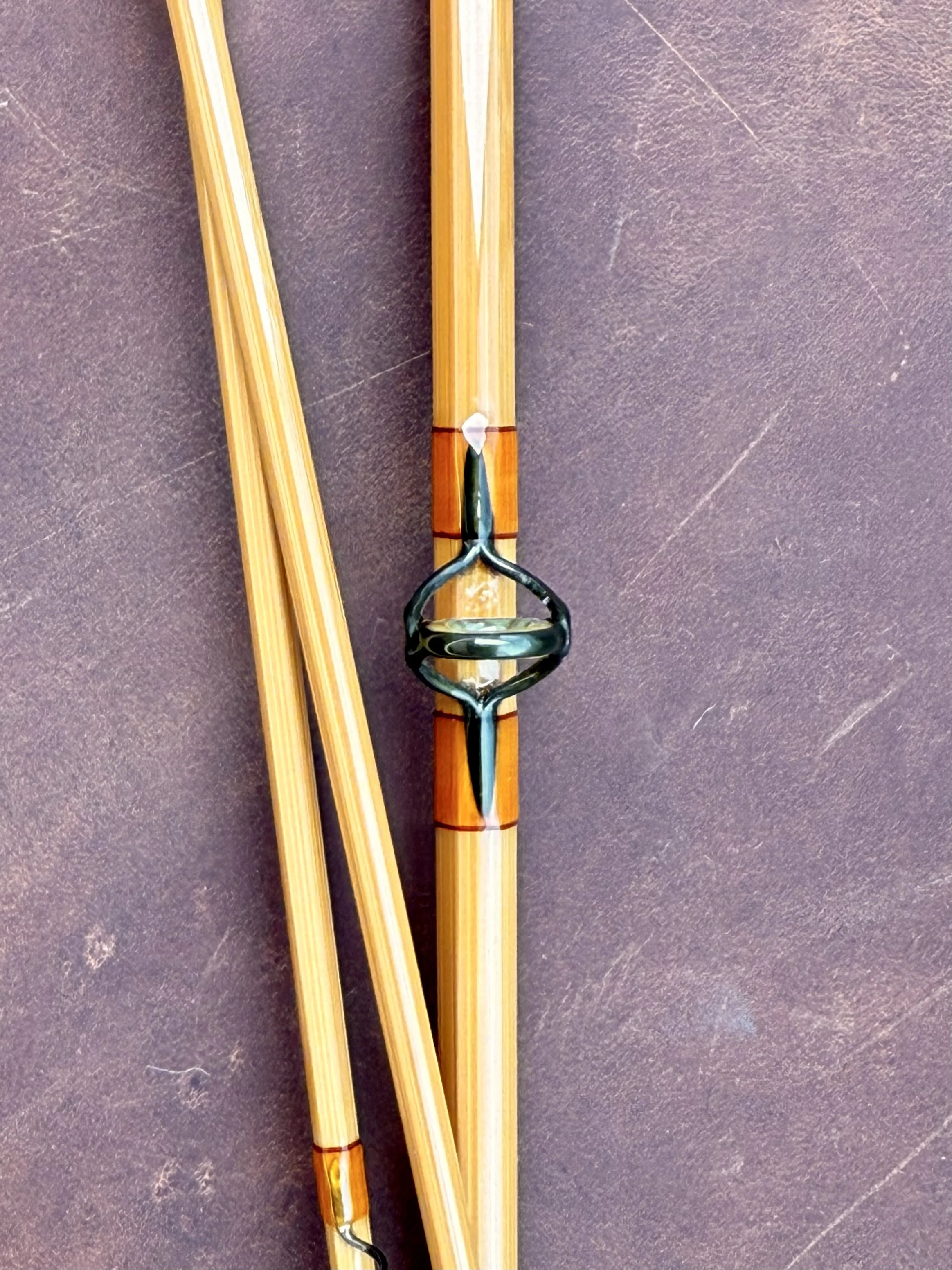 Extreme close-up of a pale, un-flamed bamboo fly rod section featuring a blued stripping guide, wrapped with burnt orange thread accented by thin dark trim bands, alongside two lighter rod sections on a leather surface.
