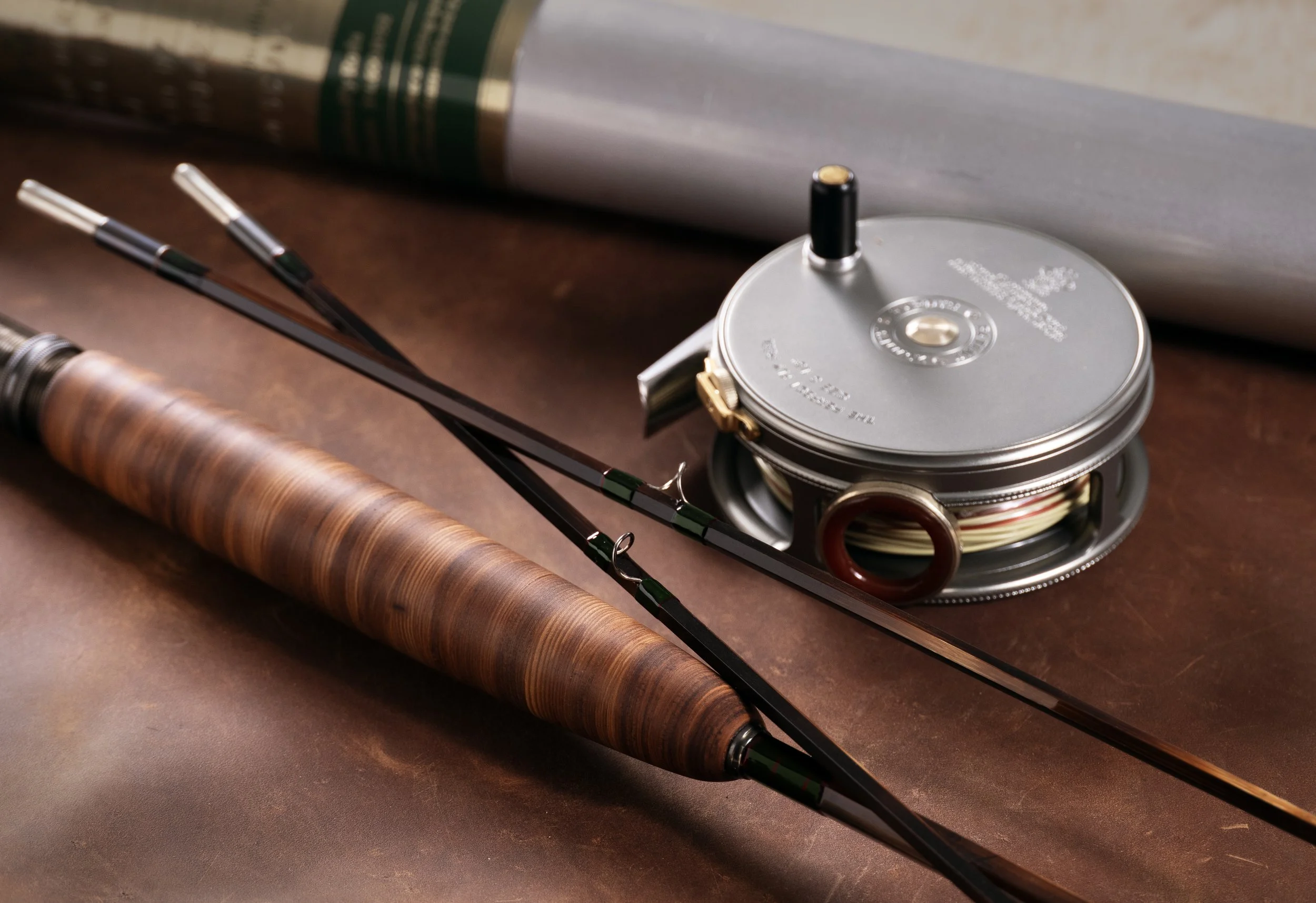 A handcrafted fly rod with a cottonwood bark grip and two tip sections laid on a brown leather surface beside a vintage-style fly reel with agate line guard loaded with fly line, with a rod tube in the background.