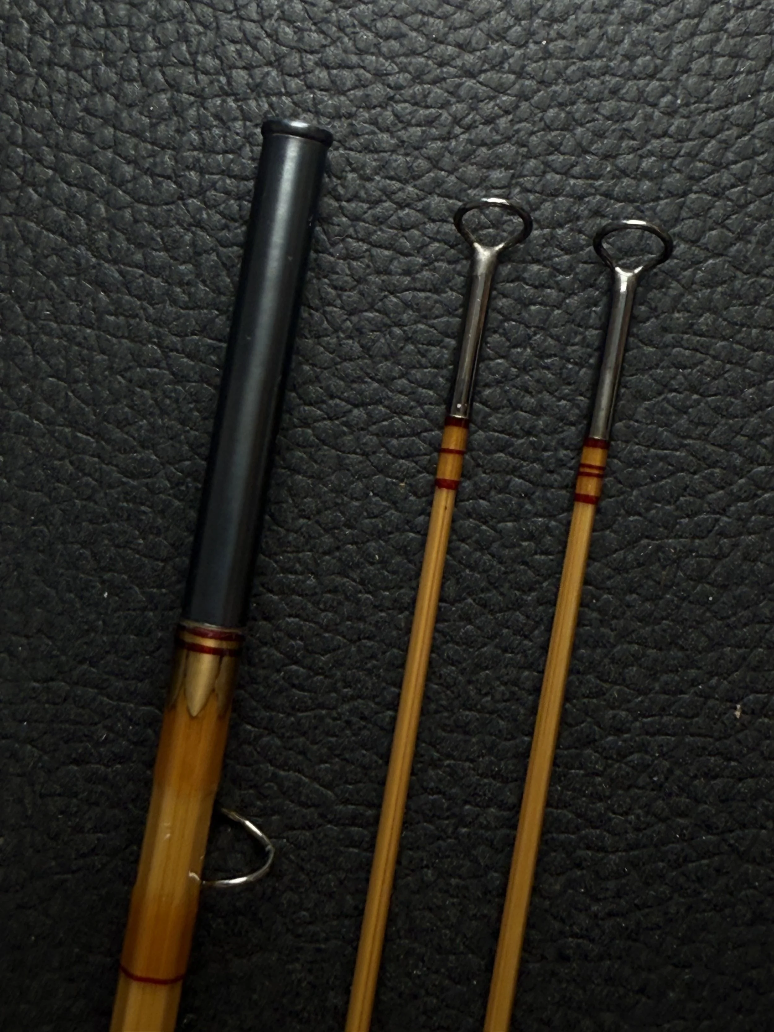 Three bamboo fly rod sections displayed against a dark leather surface, showing two  tip-top guides with loop eyes on the slender tips and antique gold thread wraps with red trim wraps at the ferrule connections, alongside a blued ferrule with crowne