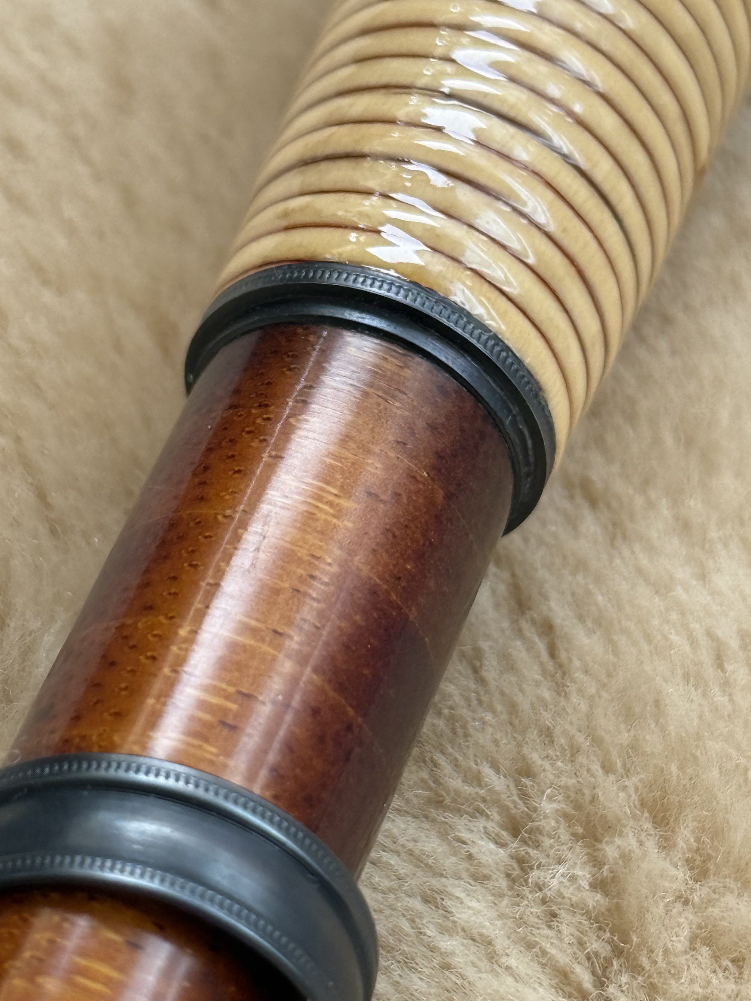 Close-up of a fly rod grip junction showing rattan grip transitioning into a flamed bamboo reel seat, separated by a knurled cork check ring, on a warm sheepskin background.