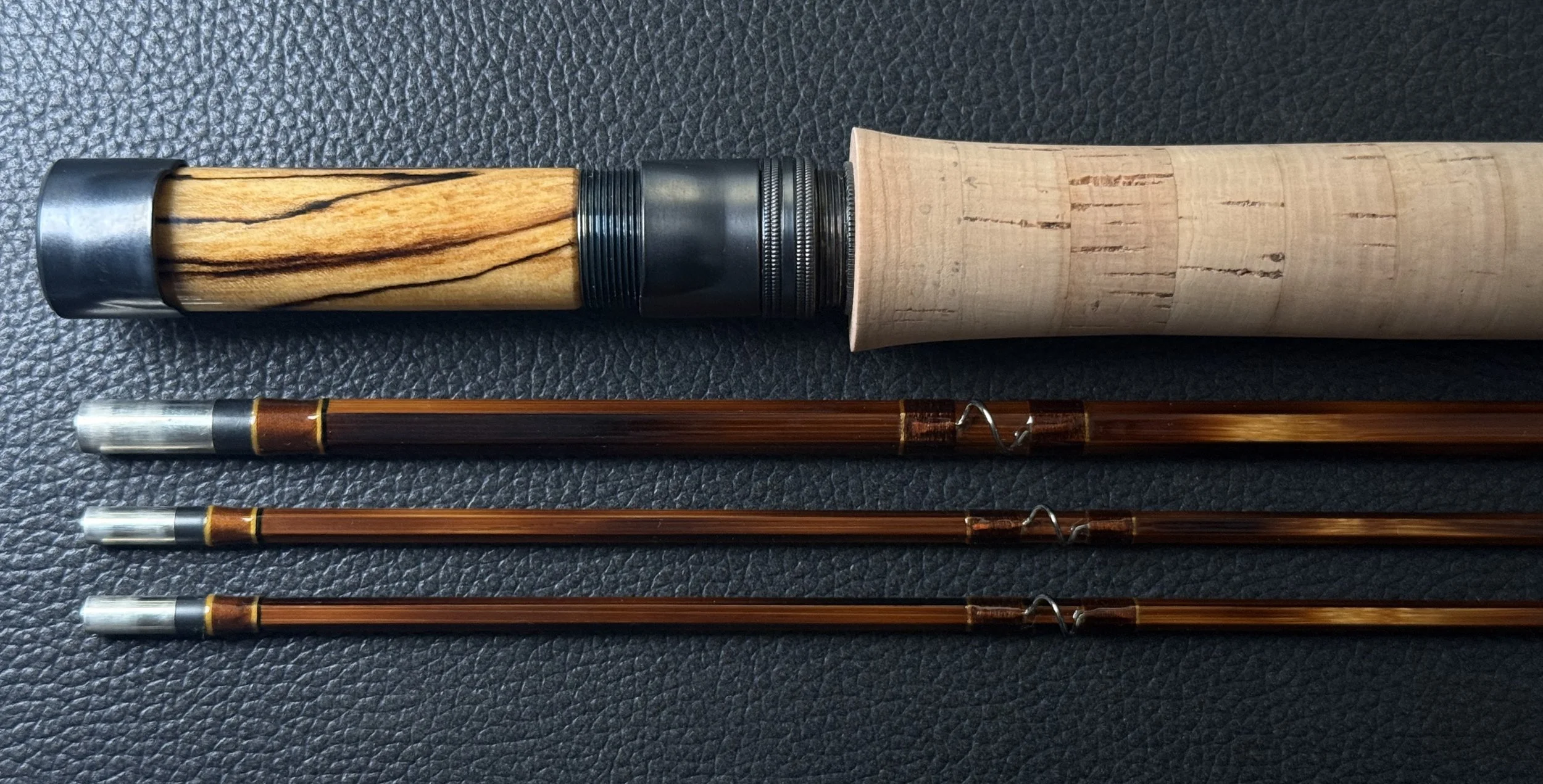 A handcrafted fly rod handle assembly featuring a striking spalted maple reel seat with blued hardware and a cork grip, displayed above three bamboo rod sections with blued ferrules, golden thread wraps, and snake guides, all on a dark leather surfac