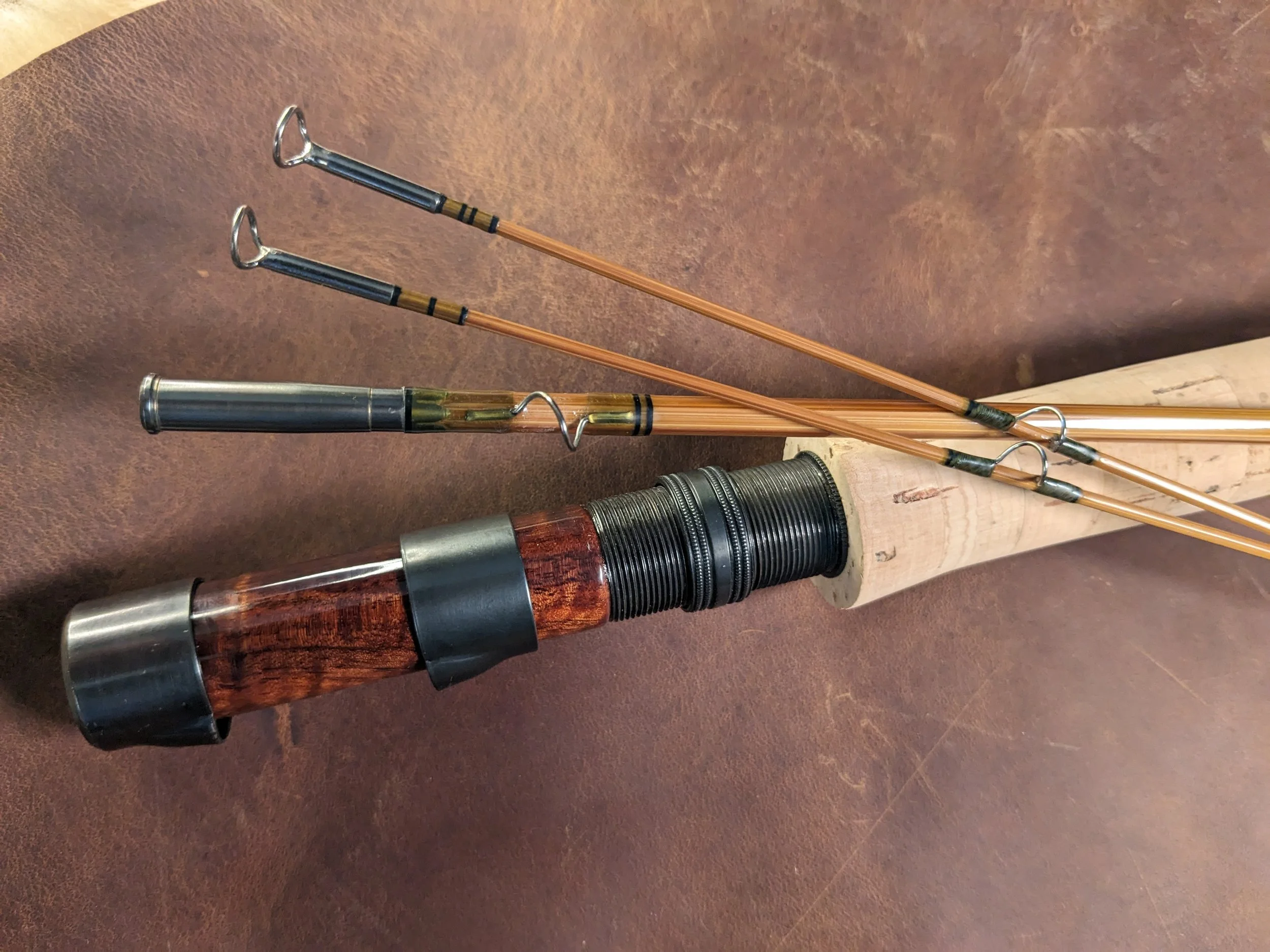 Overhead view of a handcrafted bamboo fly rod handle area featuring a richly figured curly koa wood reel seat with blued hardware and threaded locking ring, with a natural cork grip fanning to the right, and two tip sections and a mid section showing