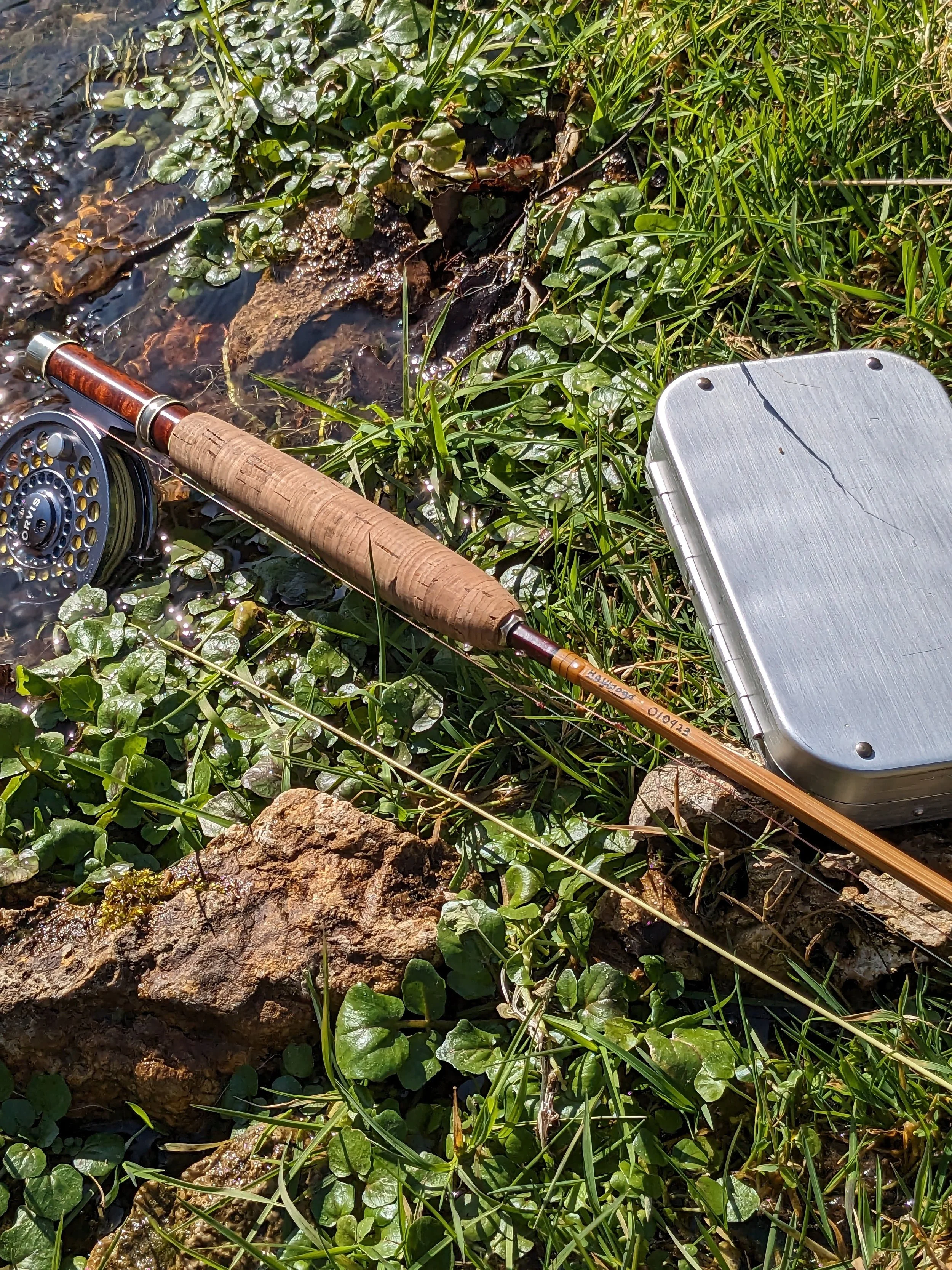 A Haywood-marked bamboo fly rod with a cork grip, burl wood reel seat, and an Orvis fly reel attached, resting on lush green streamside vegetation beside a brushed aluminum fly box and mossy rocks at the water's edge on a sunny day.